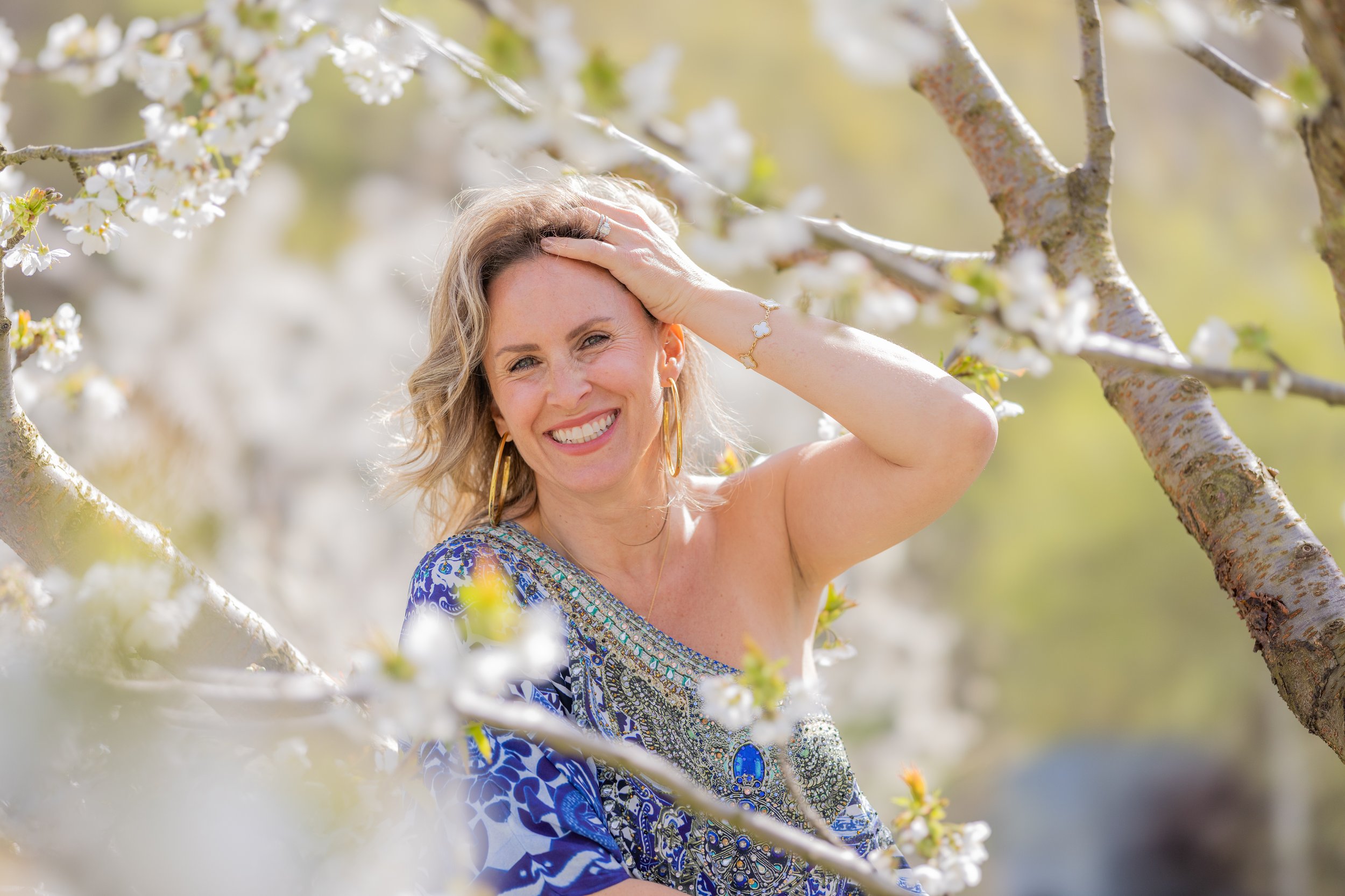 A woman with blonde wavy hair wearing a colorful patterned blue dress, large hoop earrings, and a bracelet, smiling happily in a blooming orchard of white flowers, with sunlight filtering through the trees.