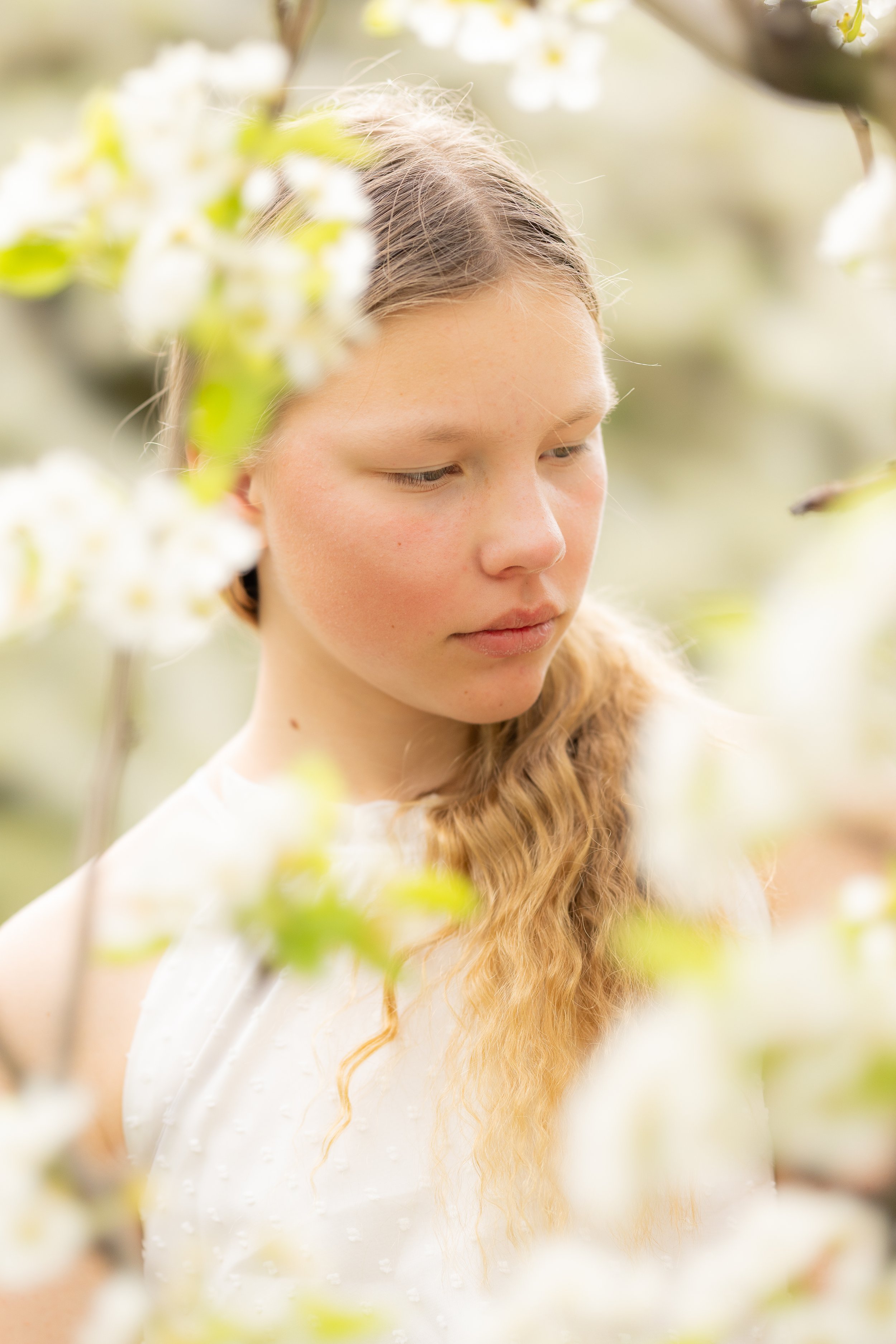 A young woman with long, wavy, reddish-blonde hair standing among blooming white flowers and looking downward thoughtfully.