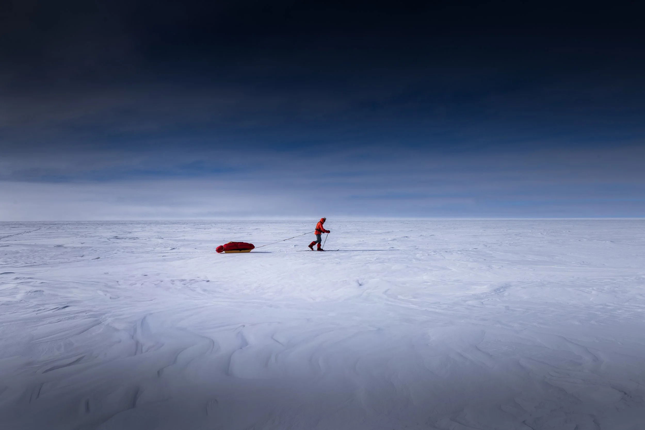 A person dressed in red and black winter gear walking across a vast, snowy, and icy landscape while pulling a red and yellow sled behind them, under a dark, cloudy sky.