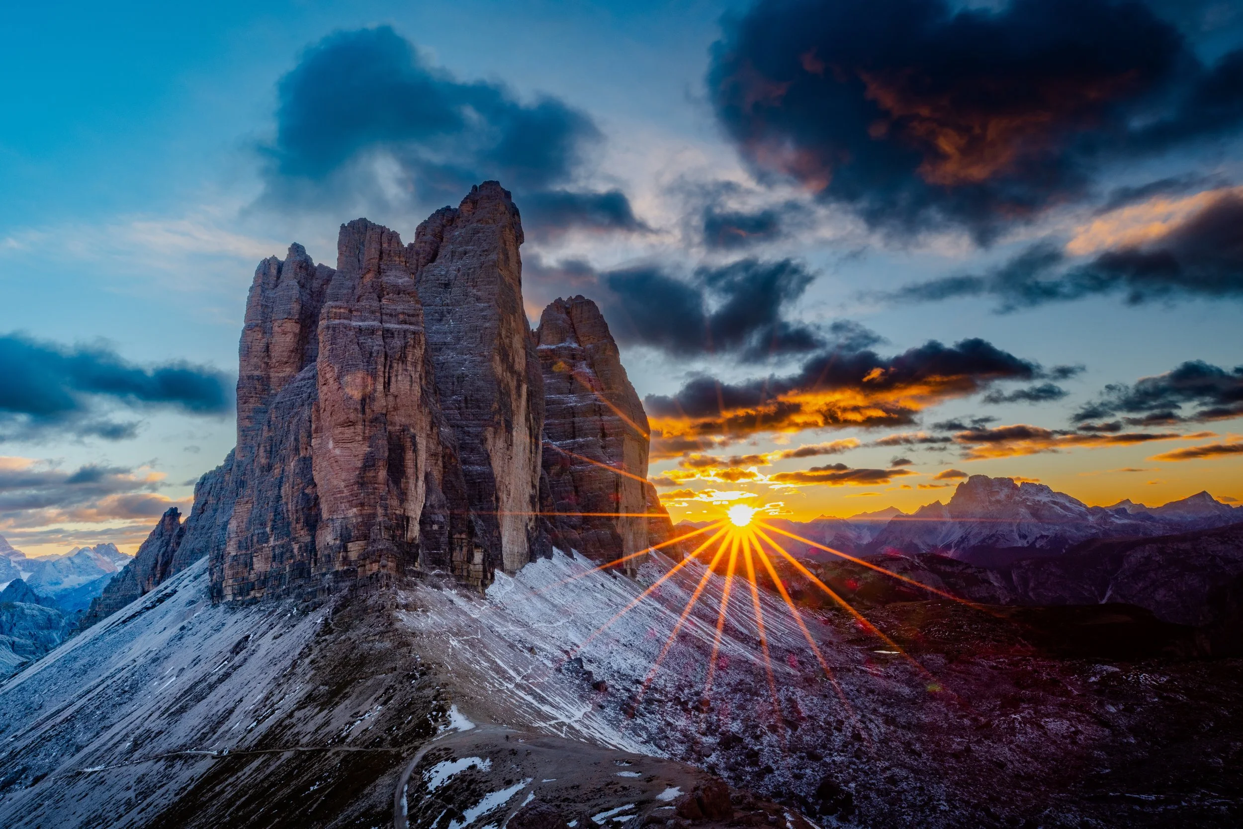 A mountain landscape at sunset with a large rocky mountain in the foreground, snow on the ground, and the sun setting behind distant mountains, partly cloudy sky.
