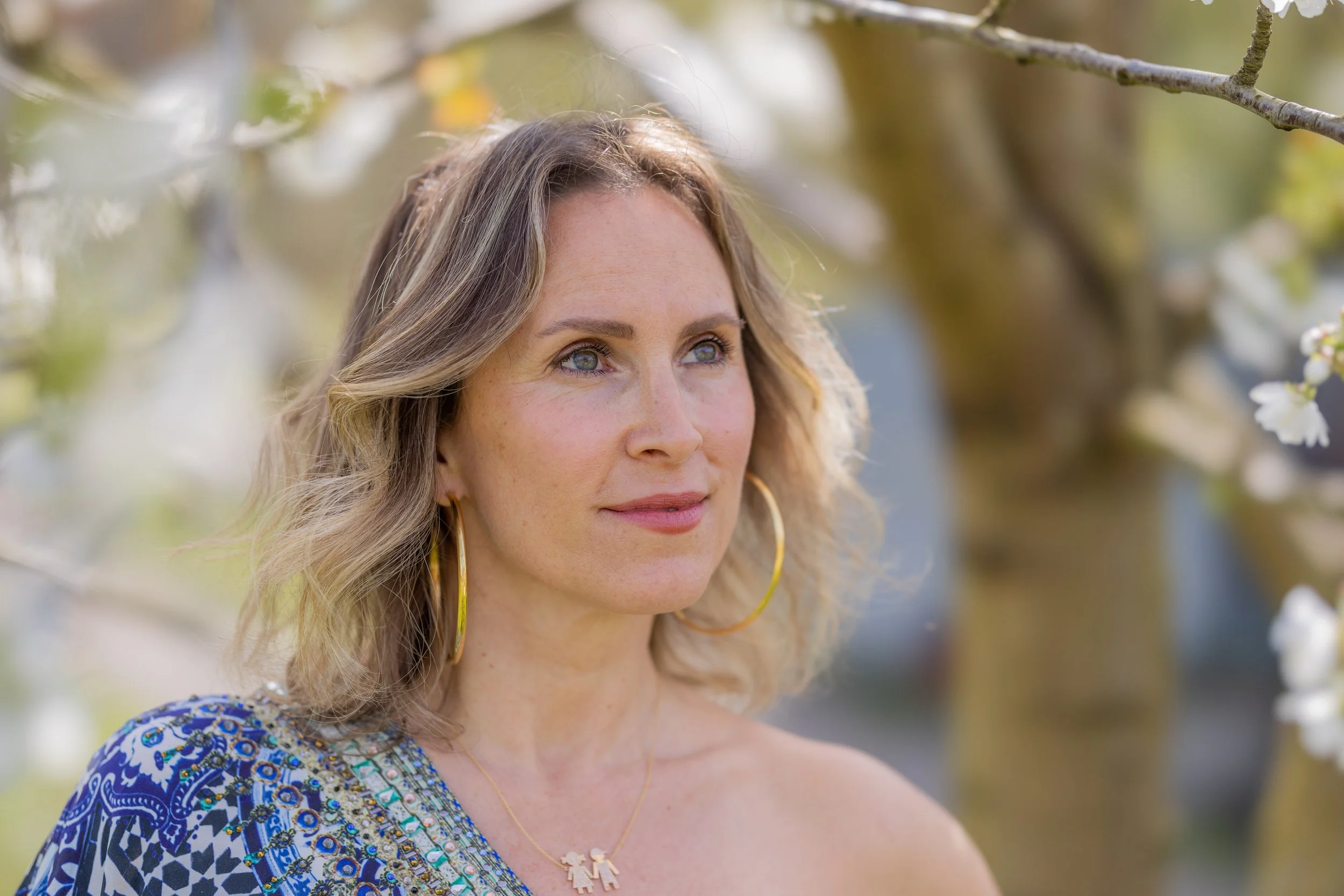 A woman with shoulder-length blonde hair looking to the side, wearing gold hoop earrings, a gold necklace, and a blue patterned off-shoulder top, outdoors with a blurred background of trees and white blossoms.