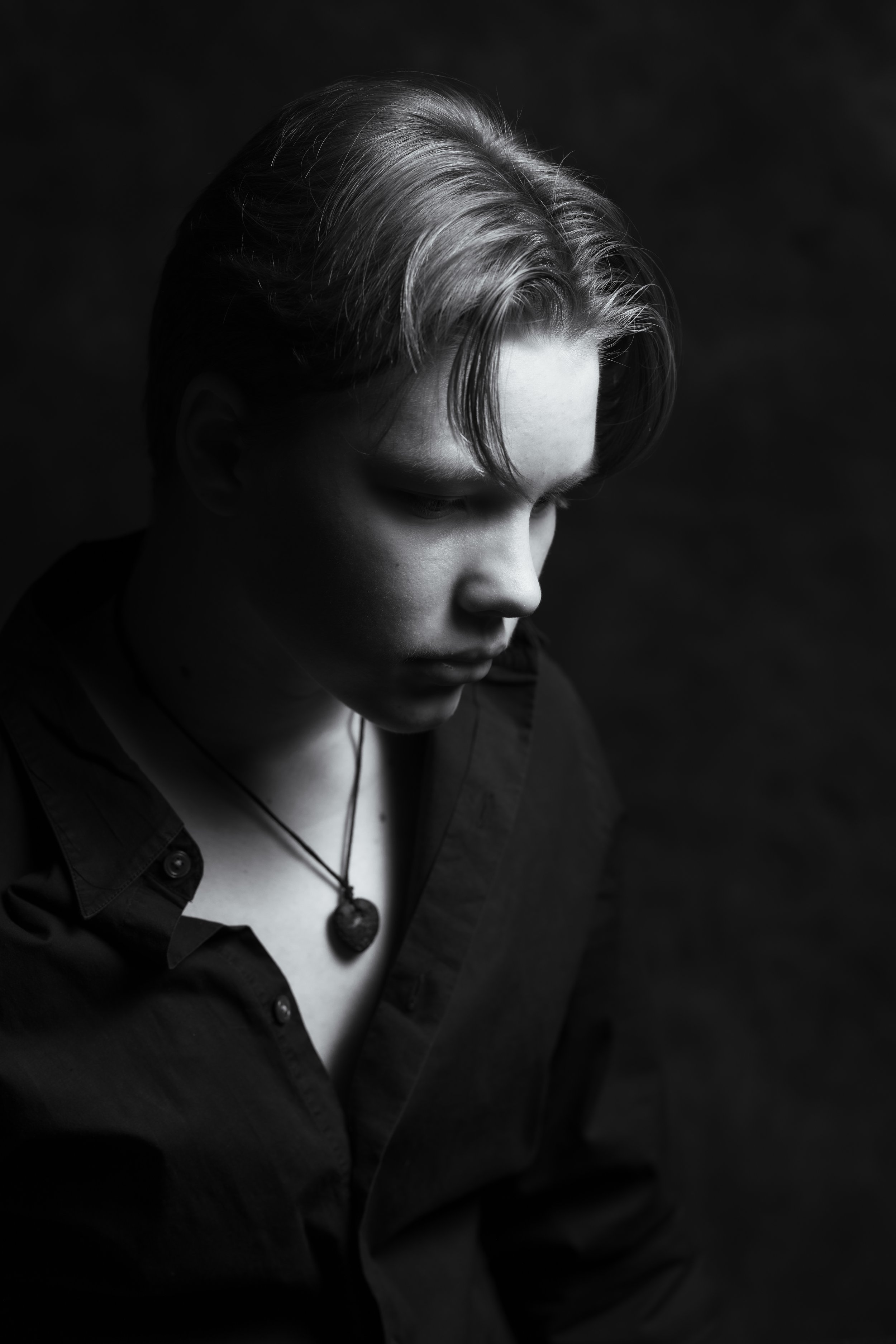 A black and white portrait of a young man with light hair looking down, wearing a dark shirt and a heart-shaped pendant necklace.