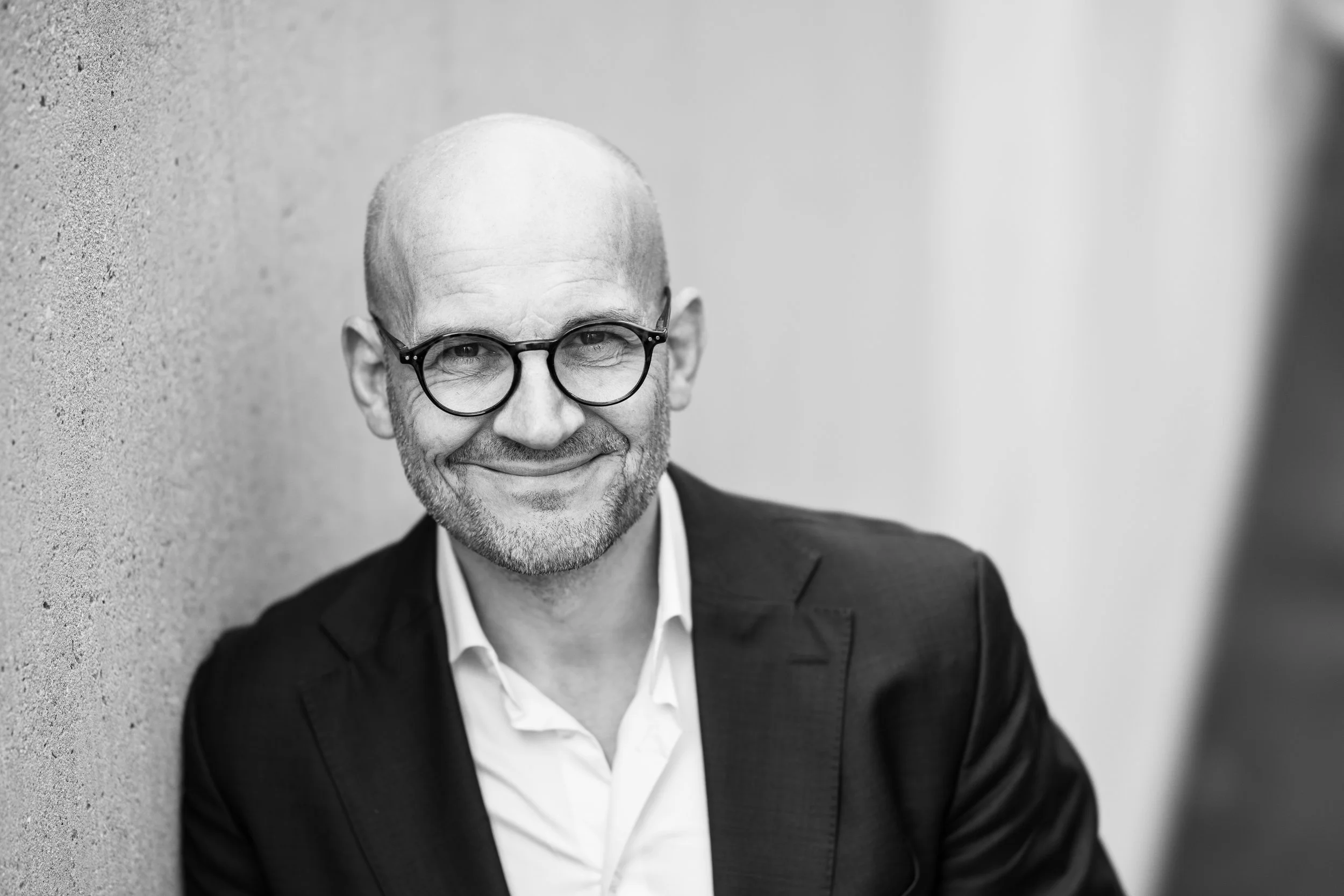 Black and white portrait of a smiling middle-aged man with glasses, wearing a suit and white shirt, leaning against a wall.