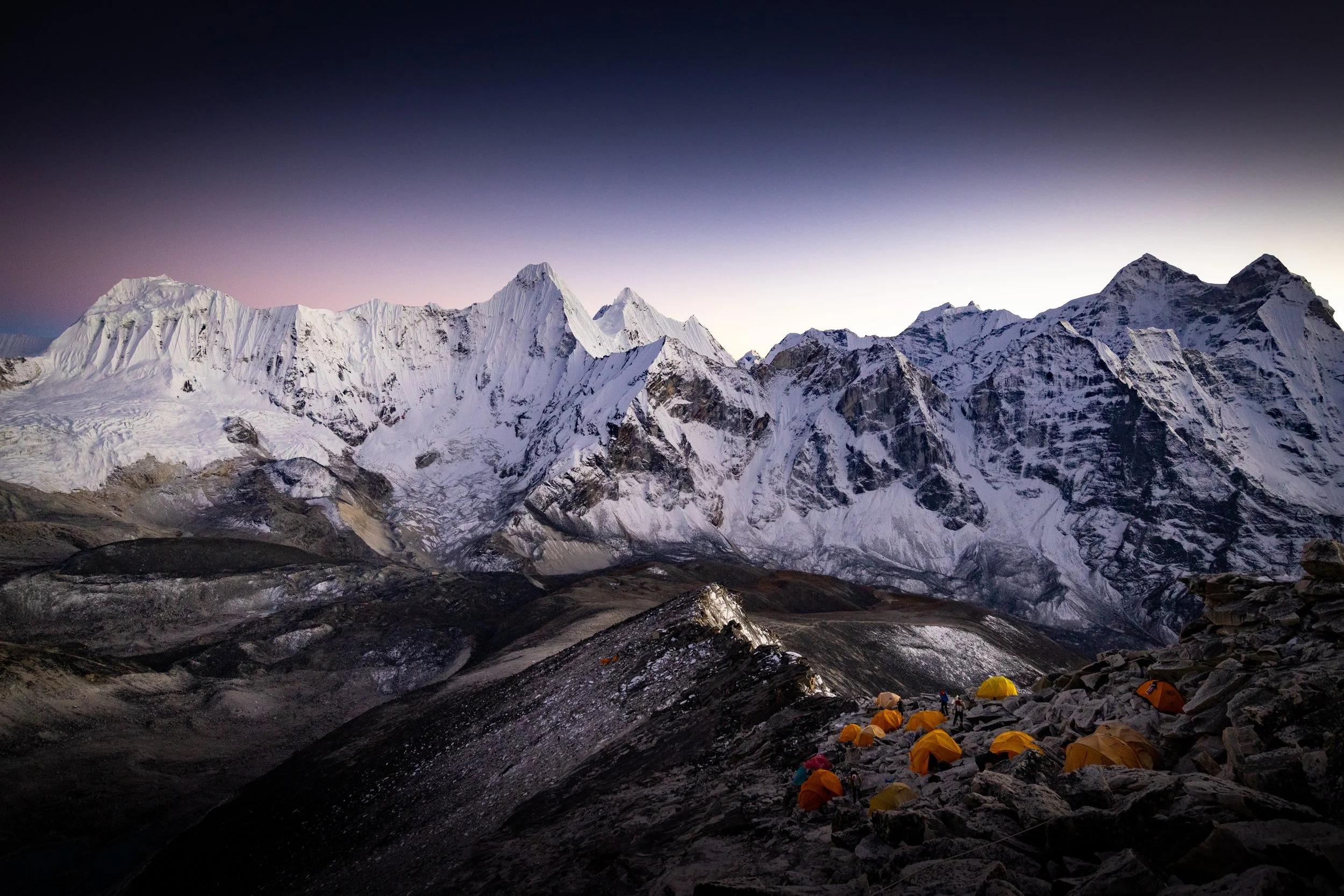 Mountains covered in snow with a campsite of yellow tents on rocky terrain in the foreground, under a twilight sky.