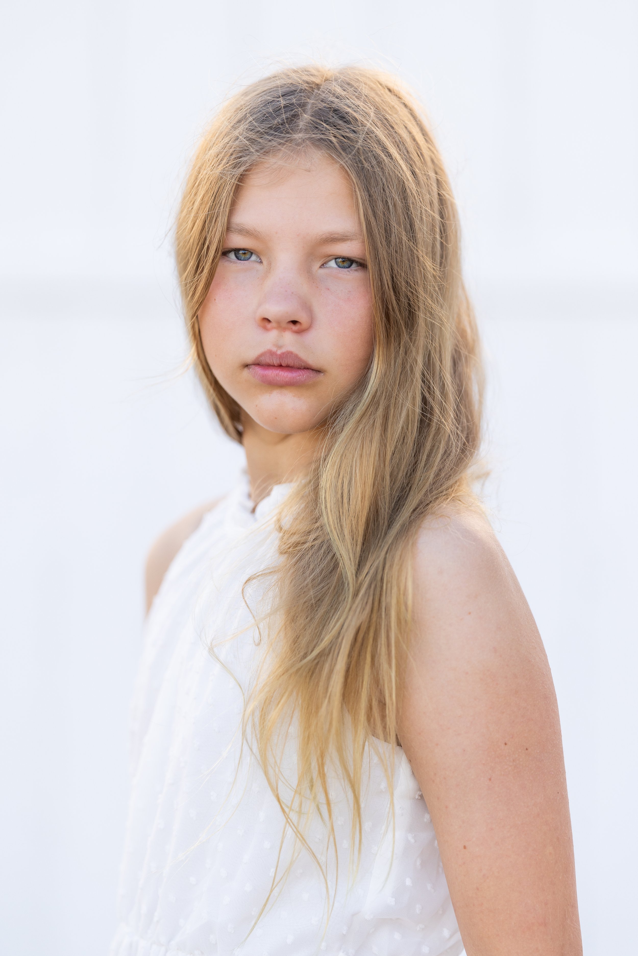 A young woman with long, wavy blonde hair and blue eyes looking at the camera against a blurred white background.