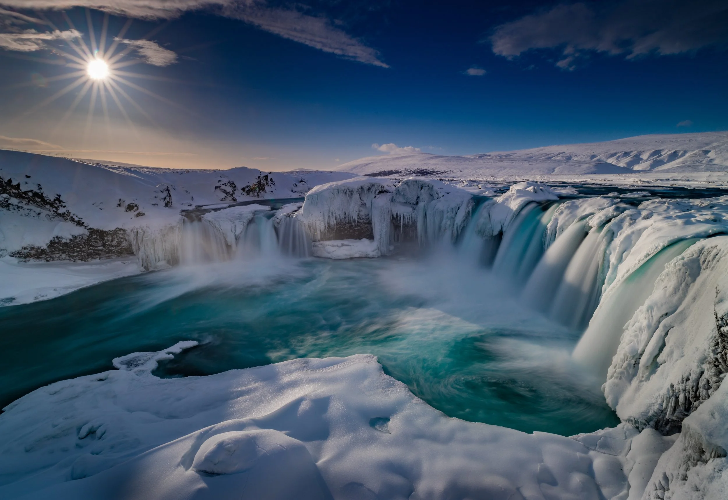 Frozen waterfall in a snowy landscape under a bright sun with a clear blue sky.