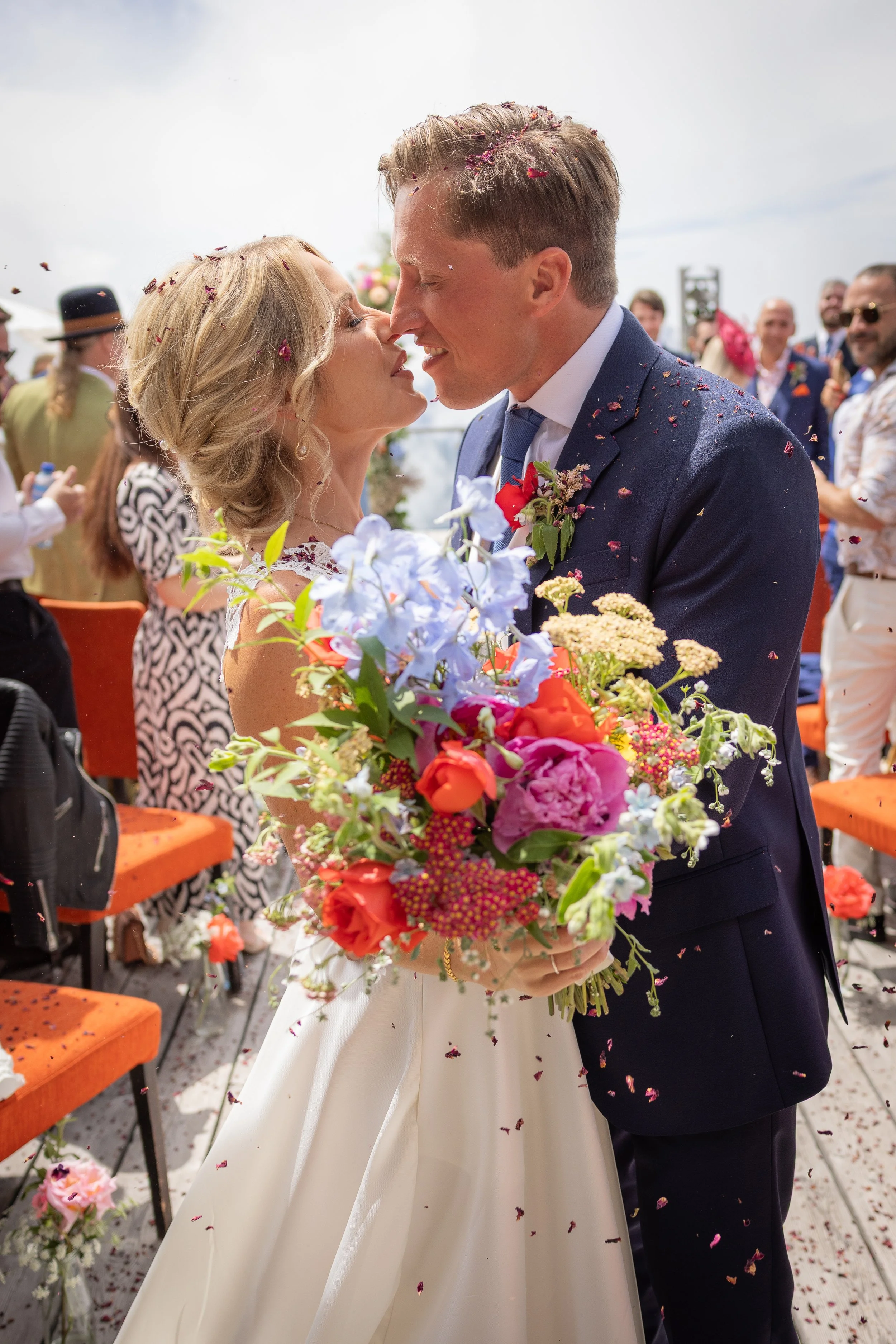 A bride and groom embrace and prepare to kiss during their wedding ceremony, surrounded by colorful flowers and guests at a wedding in Verbier Switzerland.
