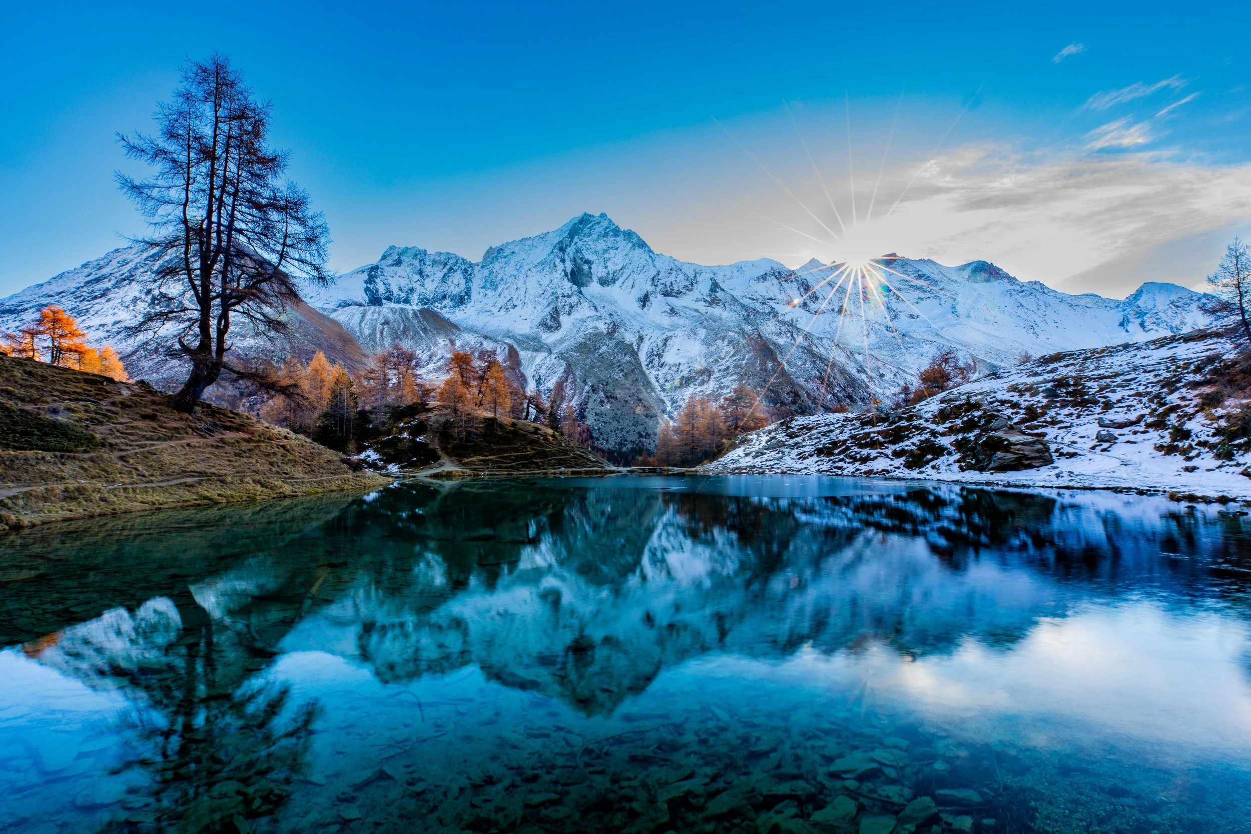 Scenic landscape of snow-capped mountains reflecting in a clear lake, with a bright sun shining in the sky and sparse trees on the hillside.