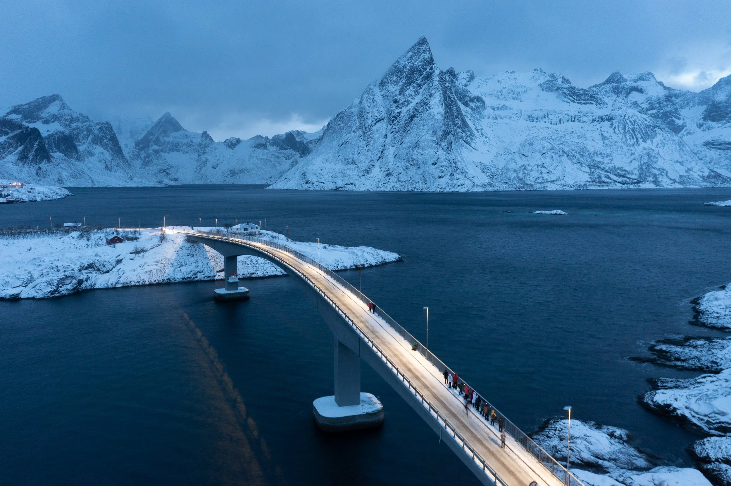 A snowy bridge over dark water with snow-covered mountains in the background at dusk.