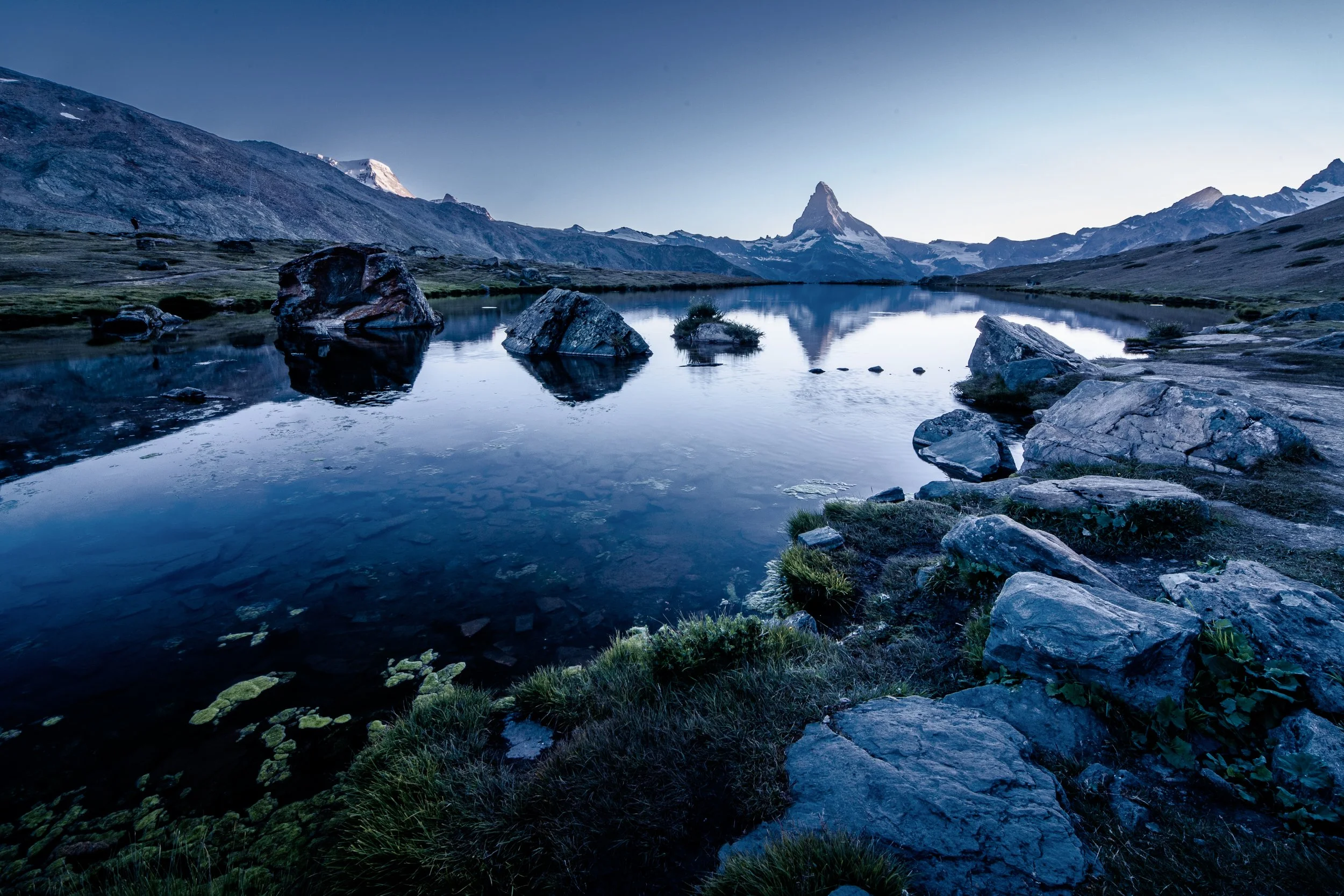 A mountain landscape featuring a calm lake with rocks and greenery in the foreground, rugged mountains in the background, and a tall mountain peak under a clear sky.
