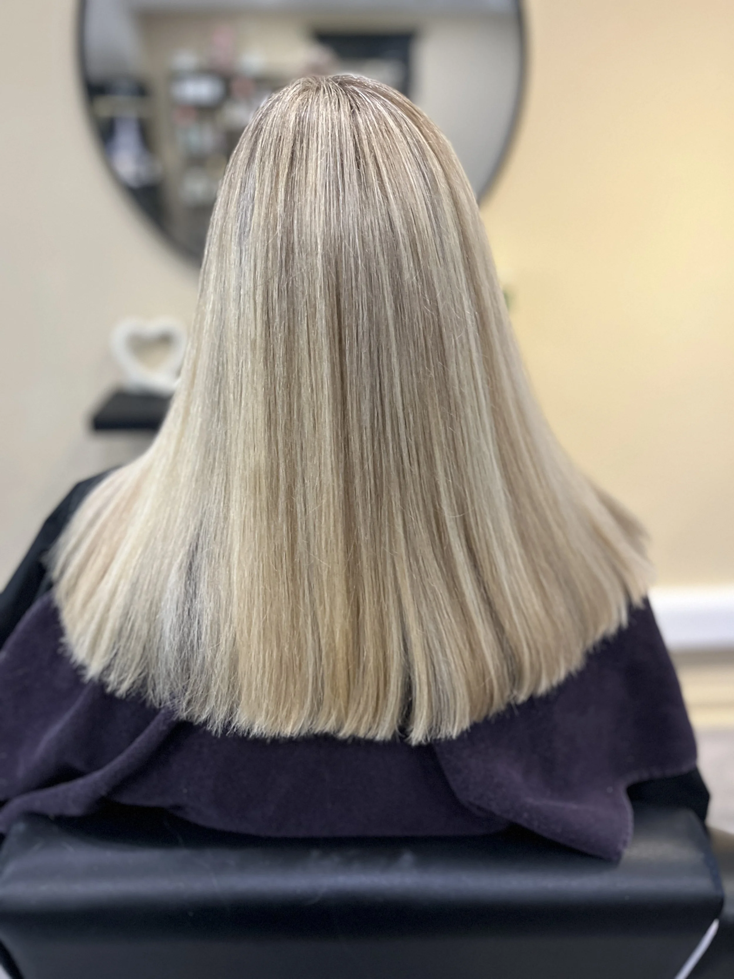 A person with shoulder-length, straight, blonde hair wearing a dark purple top, sitting near a mirror with a blurred reflection of a bedroom or salon background.