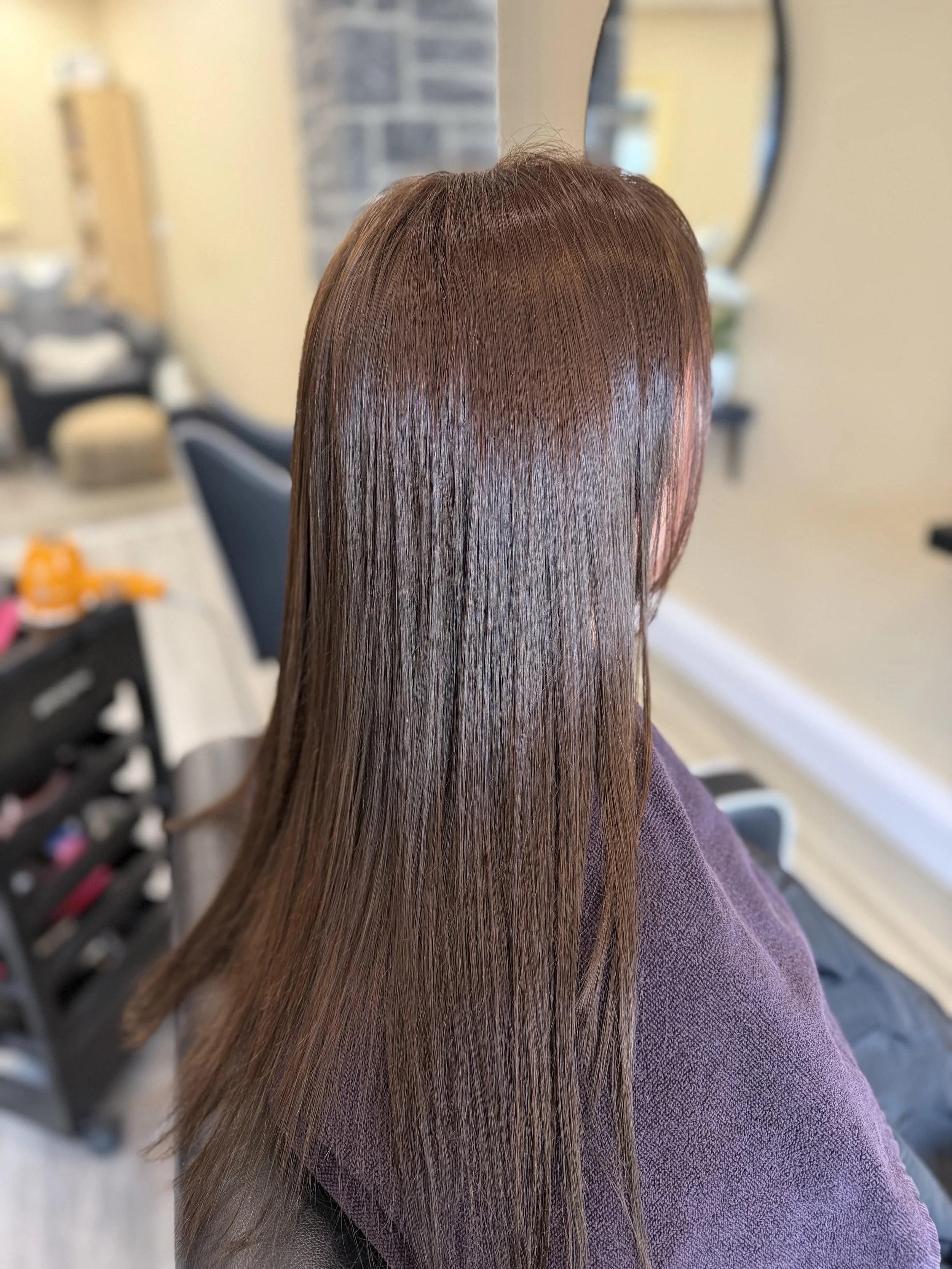 Close-up of a person with long, straight, shiny, brown hair, sitting in a hair salon with a mirror in the background.