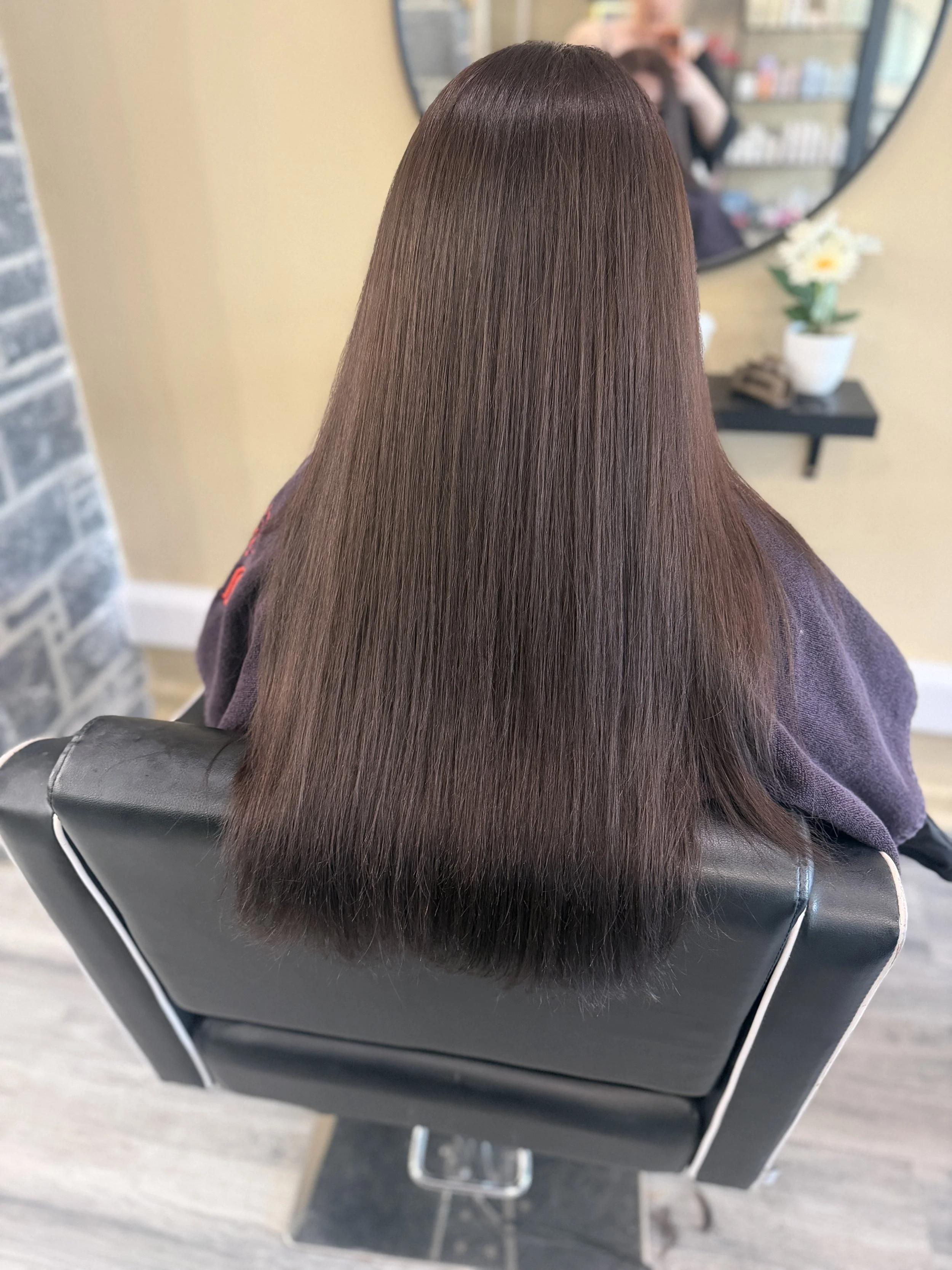 Back view of a woman with long, straight, shiny brown hair sitting in a salon chair in a hair salon, with a large mirror and a vase with white flowers in the background.