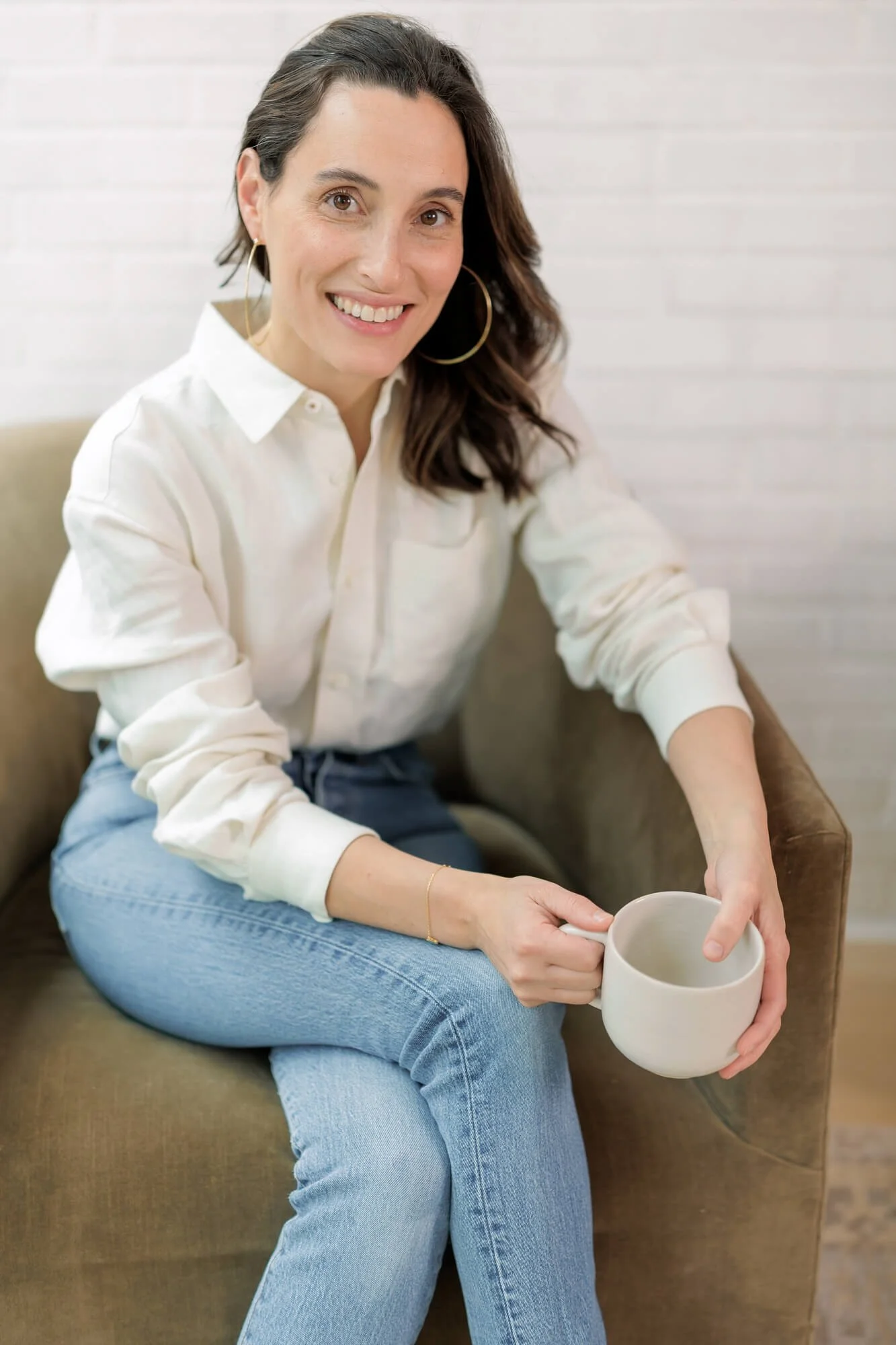 A woman with dark hair, wearing hoop earrings and a white shirt, sitting on a brown sofa and holding a beige mug in her hand, smiling at the camera in a cozy, light-colored room.