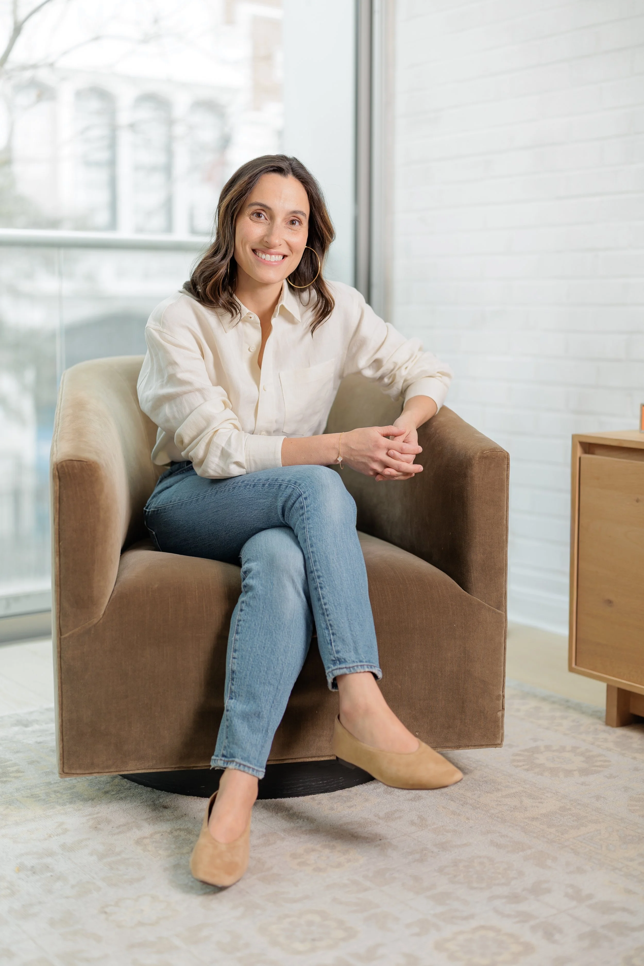 A woman smiling and sitting on a brown armchair in a bright room with large windows.