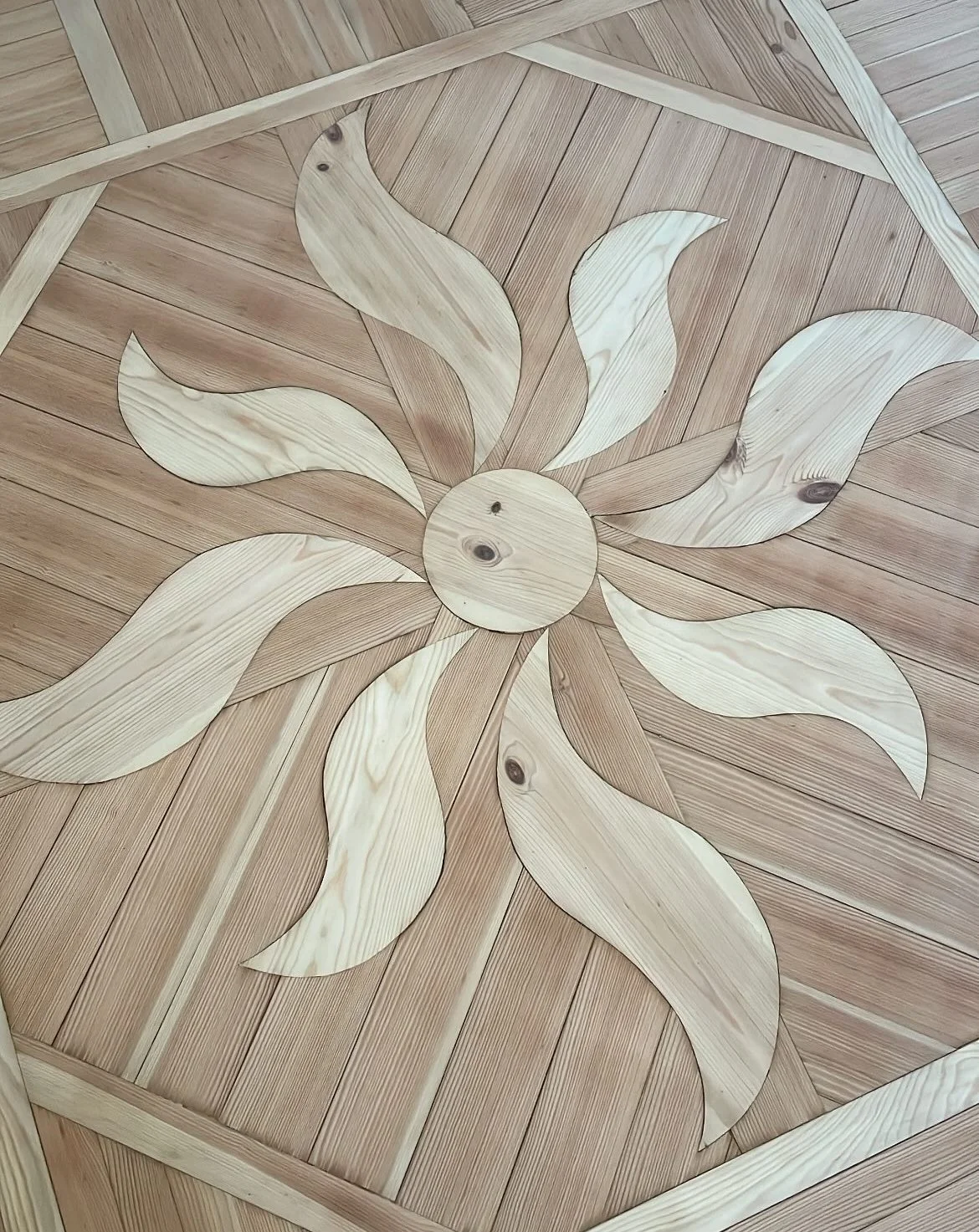 Wooden floor with inlaid design of a large flower with curved petals, made from lighter wood contrasting with the darker wood background.
