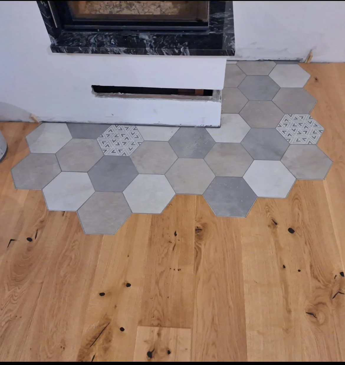 Delno tlakovana tla okoli kamina s šesterokotnimi ploščicami, nekatere z geometrijskimi vzorci, na lesenih tleh.