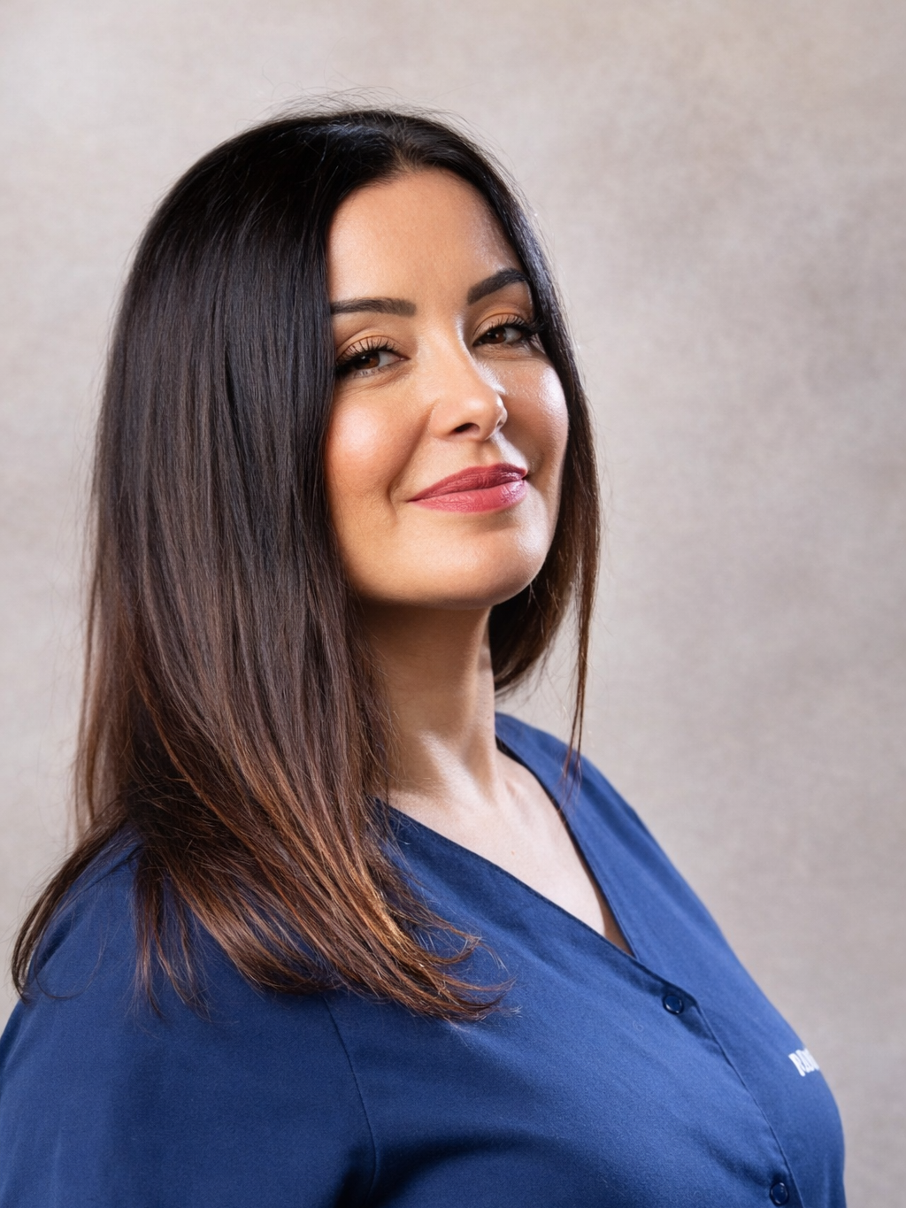 Portrait d'une femme avec des cheveux bruns longs, portant une chemise bleue, souriant légèrement, contre un fond neutre.