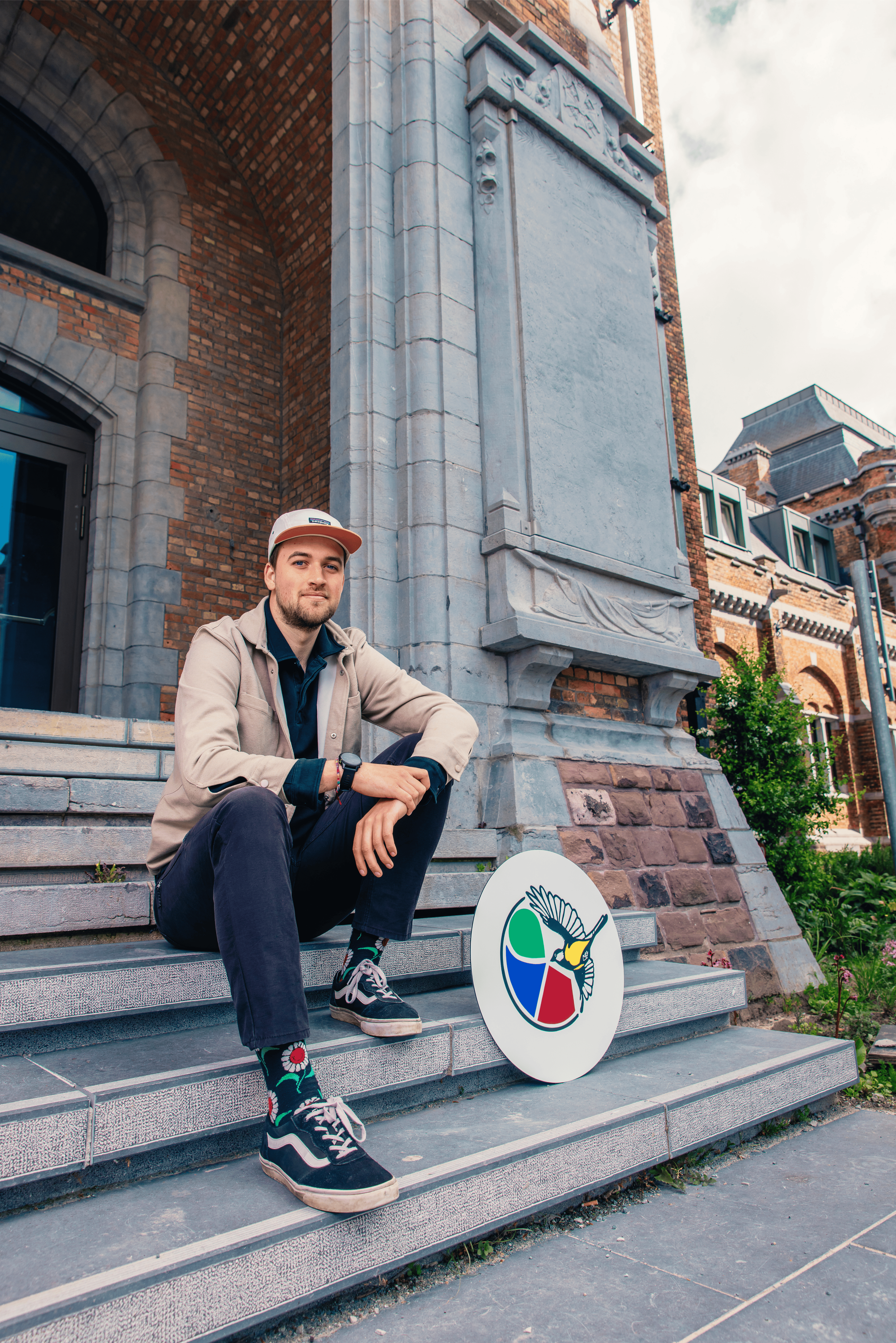 Een man in een beige jas en een witte pet zit op de trappen voor een oud bakstenen gebouw. Hij draagt kleurrijke sokken en sneakers en kijkt glimlachend naar de camera. Naast hem ligt een rond wit bord met een kleurrijk logo van een adviesbureau.