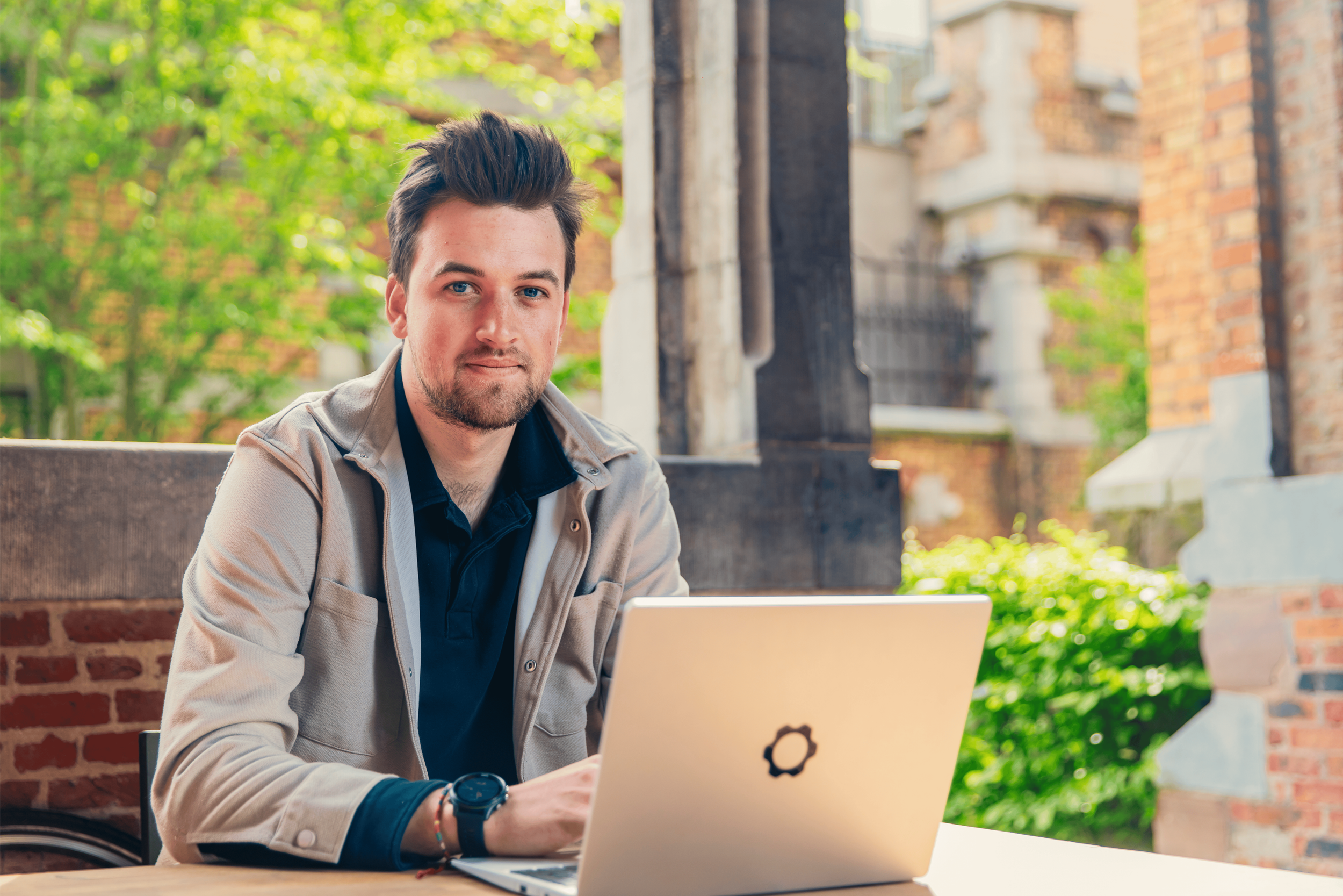 Man werkt aan een laptop op een terras met groene bomen en bakstenen muren op de achtergrond.