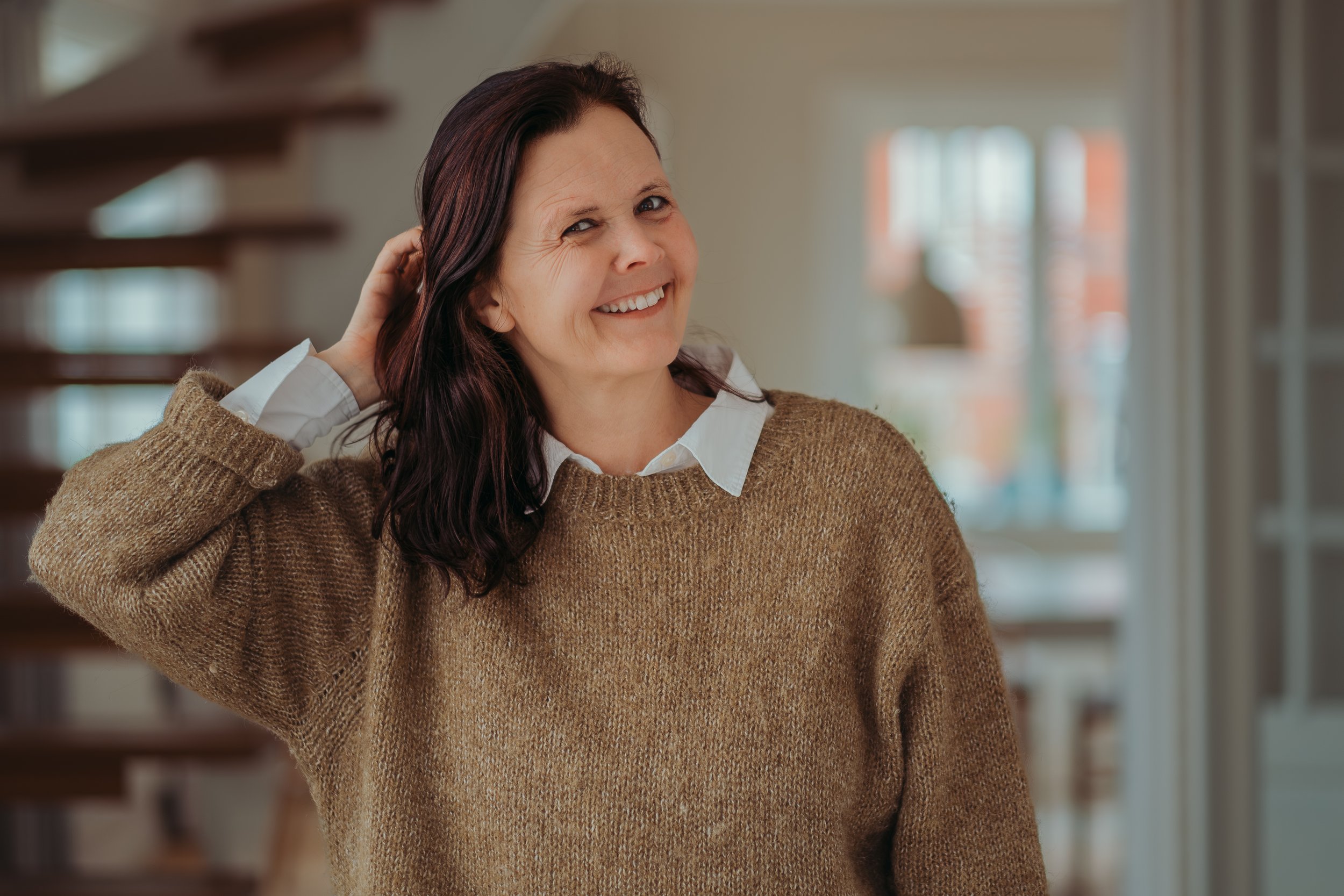 Vrouw met donker haar knuffelend haar hoofd achter haar oor, glimlachend in een huis met trappen en ramen.