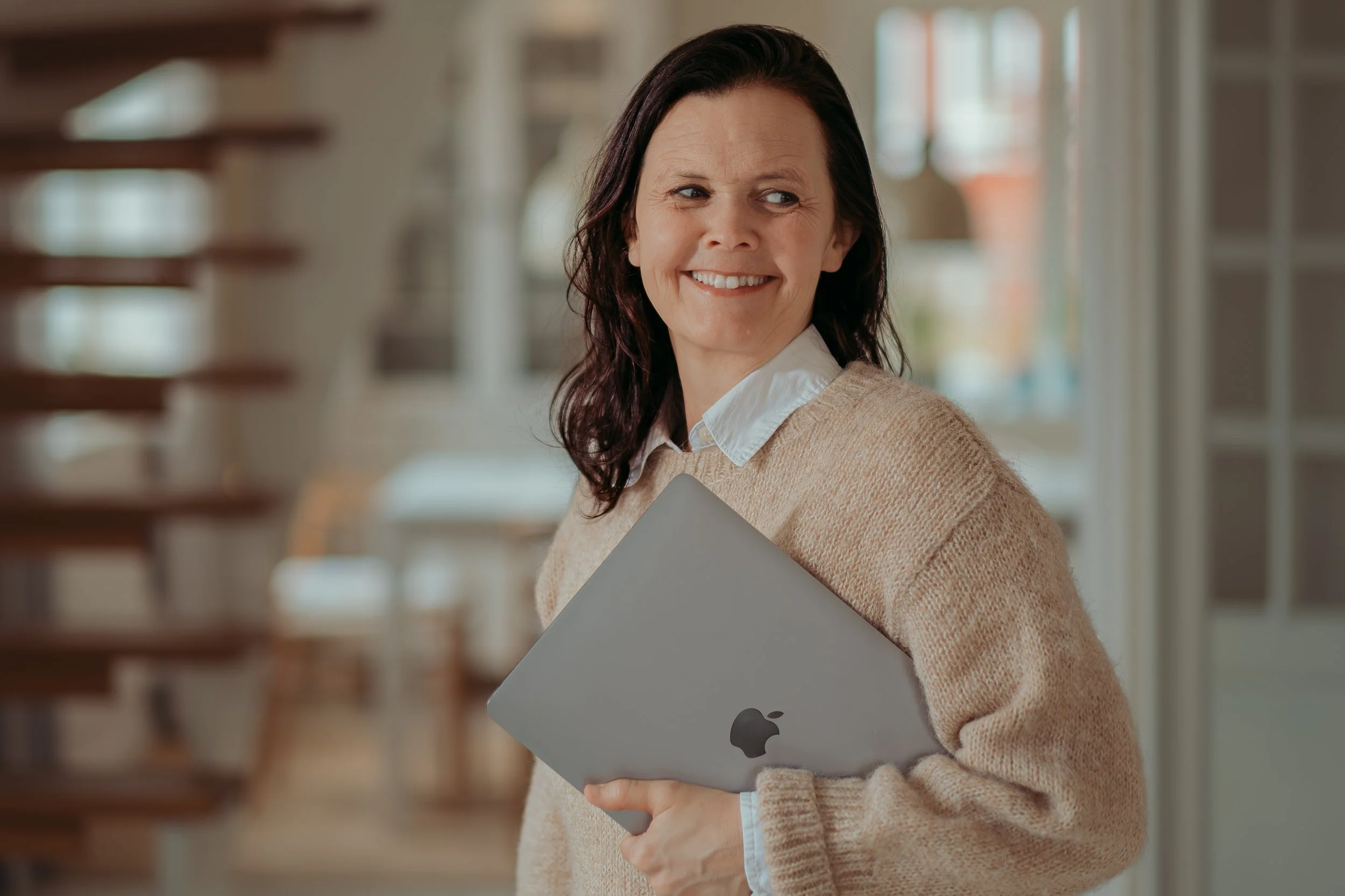 Vrolijke vrouw met bruin haar houdt een MacBook in haar rechterarm in een huis met veel natuurlijk licht, houten trappen en witte muren.