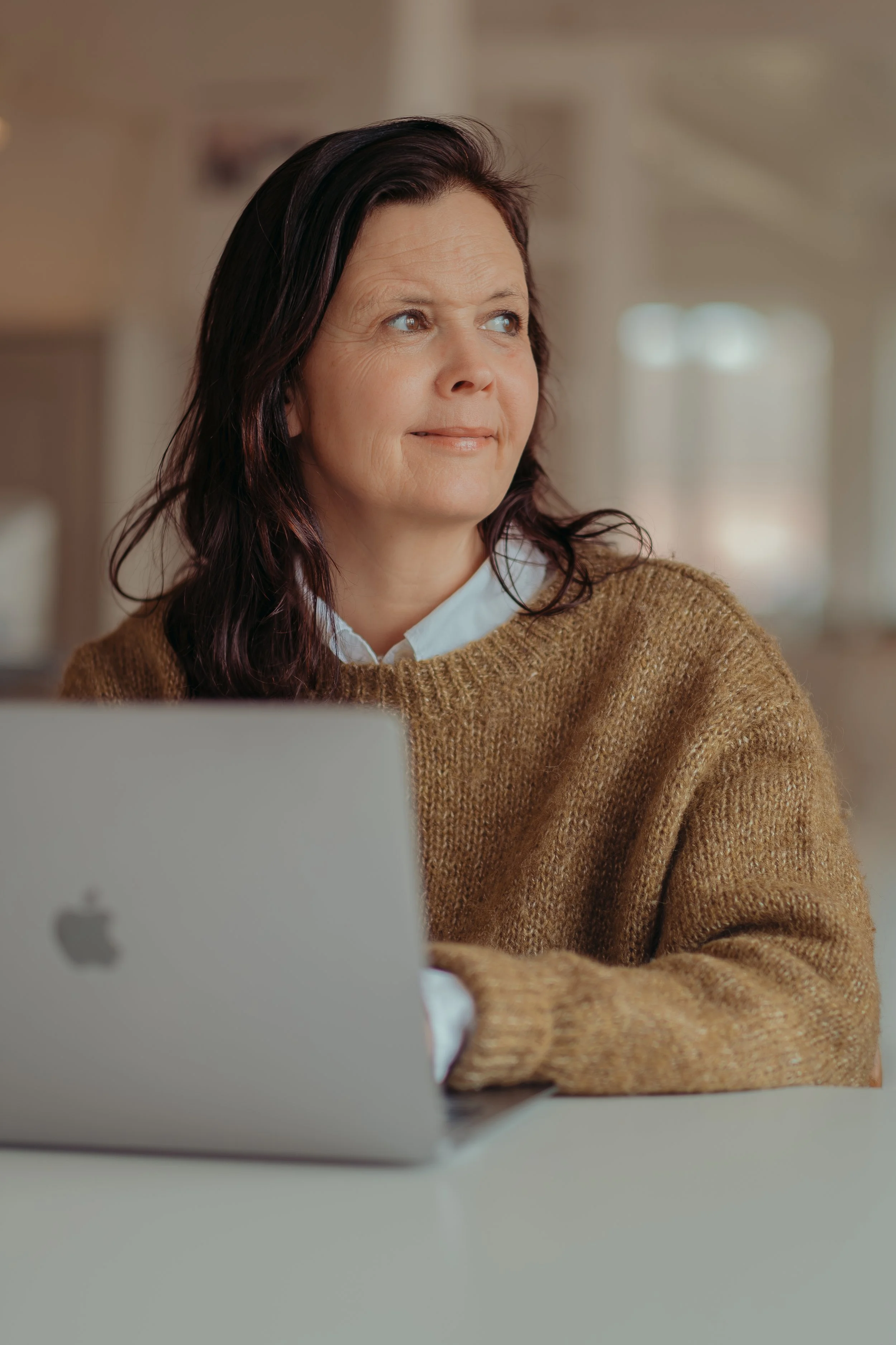 Vrouw met donkerbruin haar en een bruin trui zit aan een tafel met een zilveren MacBook, binnen- of buiten, met een relaxte uitstraling.