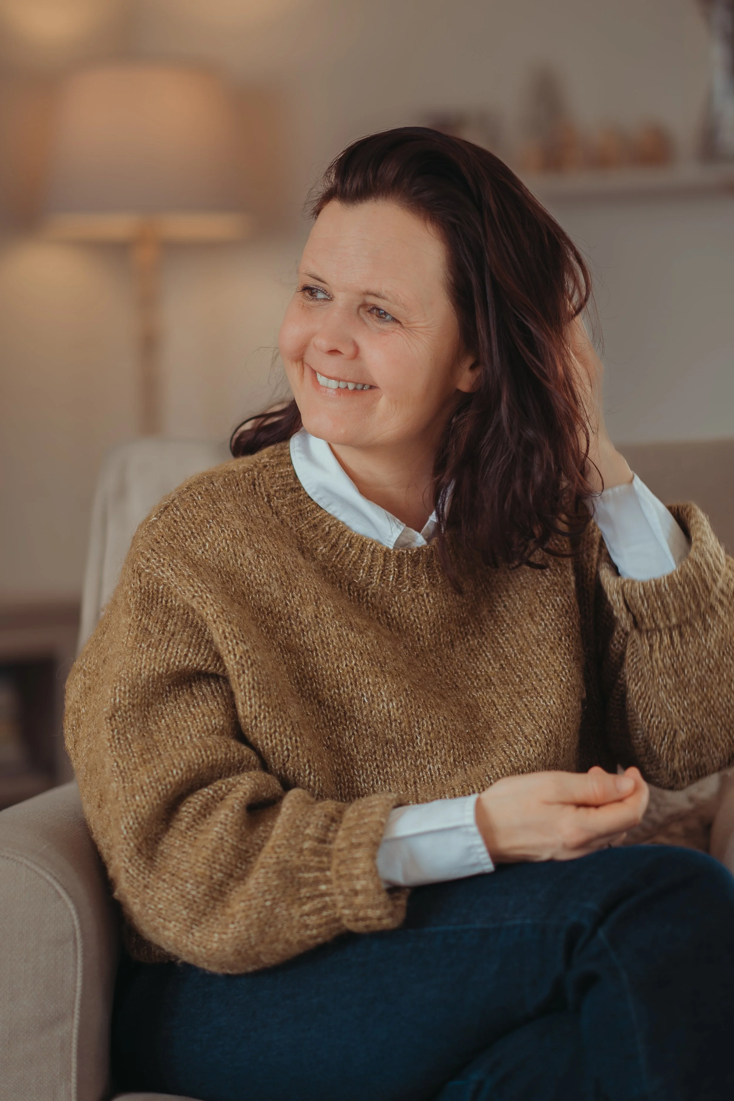 Een vrouw met donkerbruin haar, zittend op een bank, glimlacht en kijkt naar links, in een gezellige woonkamer met een zachte lamp op de achtergrond.