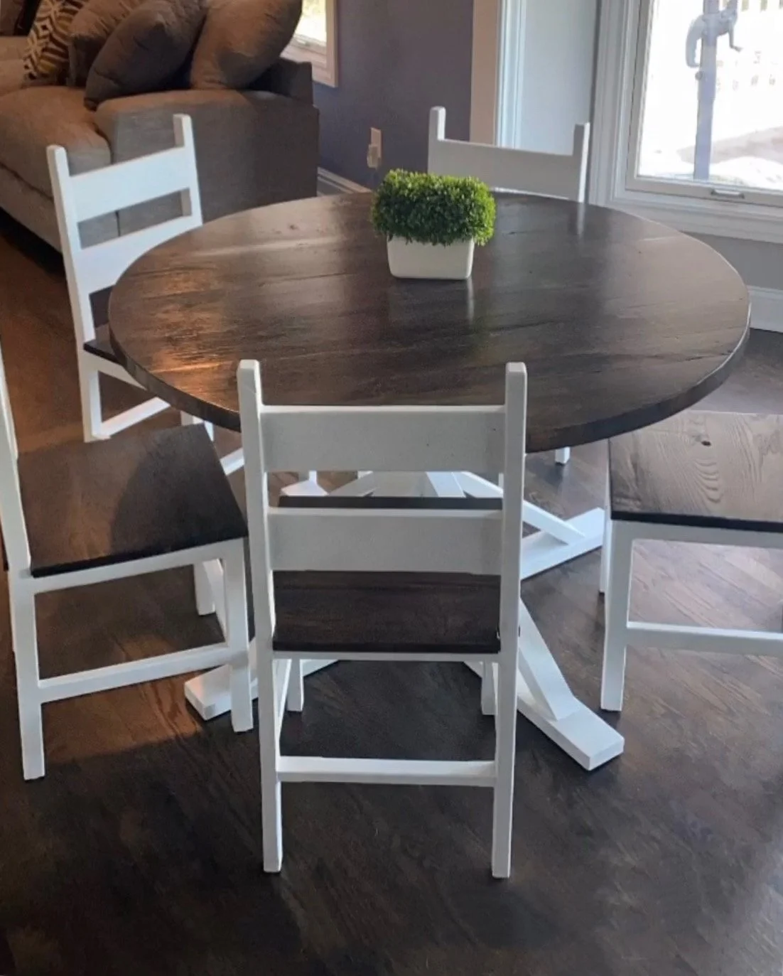 Wooden dining table with a small green potted plant on top, surrounded by four white chairs, in a room near a window.