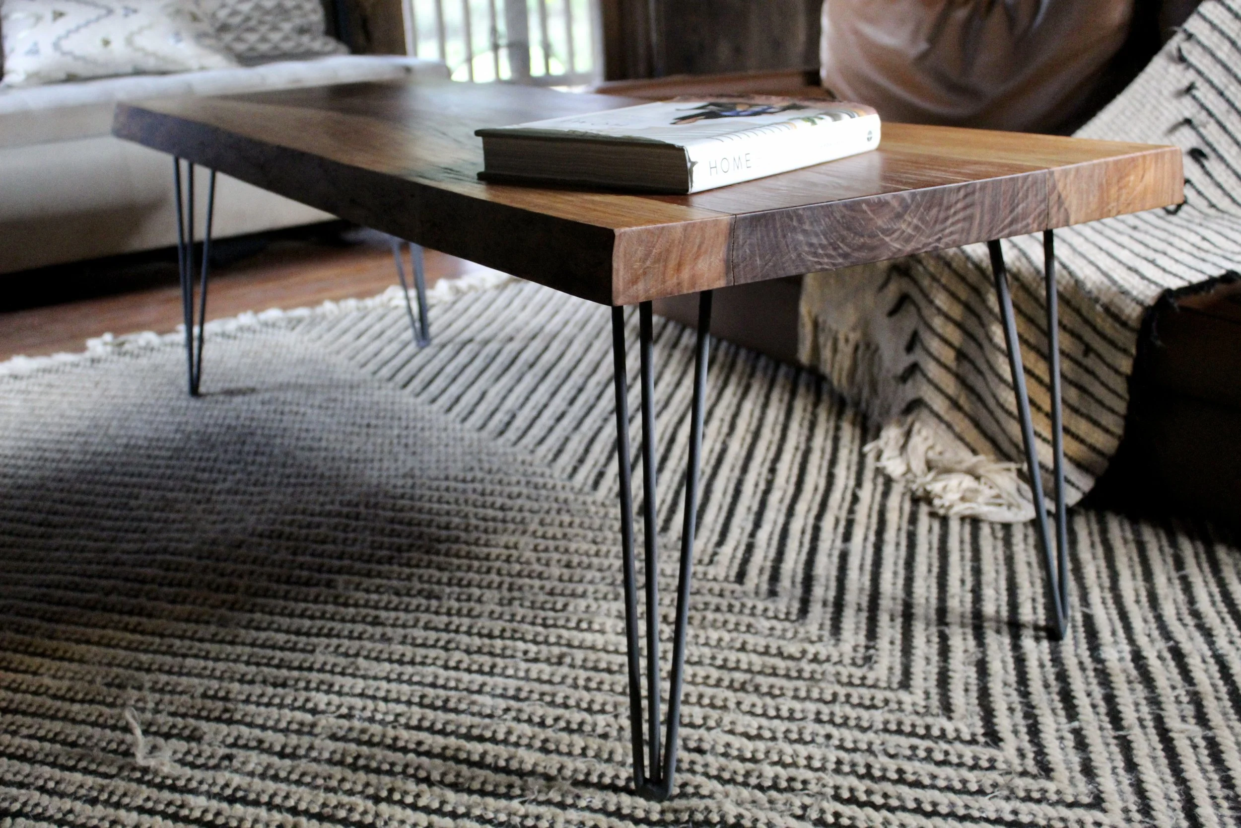 Close-up of a wooden coffee table with black metal hairpin legs, situated on a textured black and white striped rug in a cozy living room.