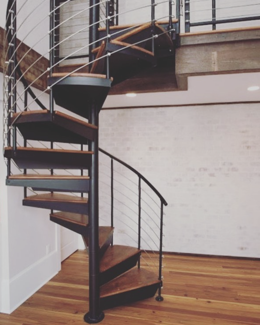 A modern indoor spiral staircase with wooden steps, black metal railings, and a curved design, leading to an upper level with a wood and metal railing, set against a white brick wall and hardwood flooring.