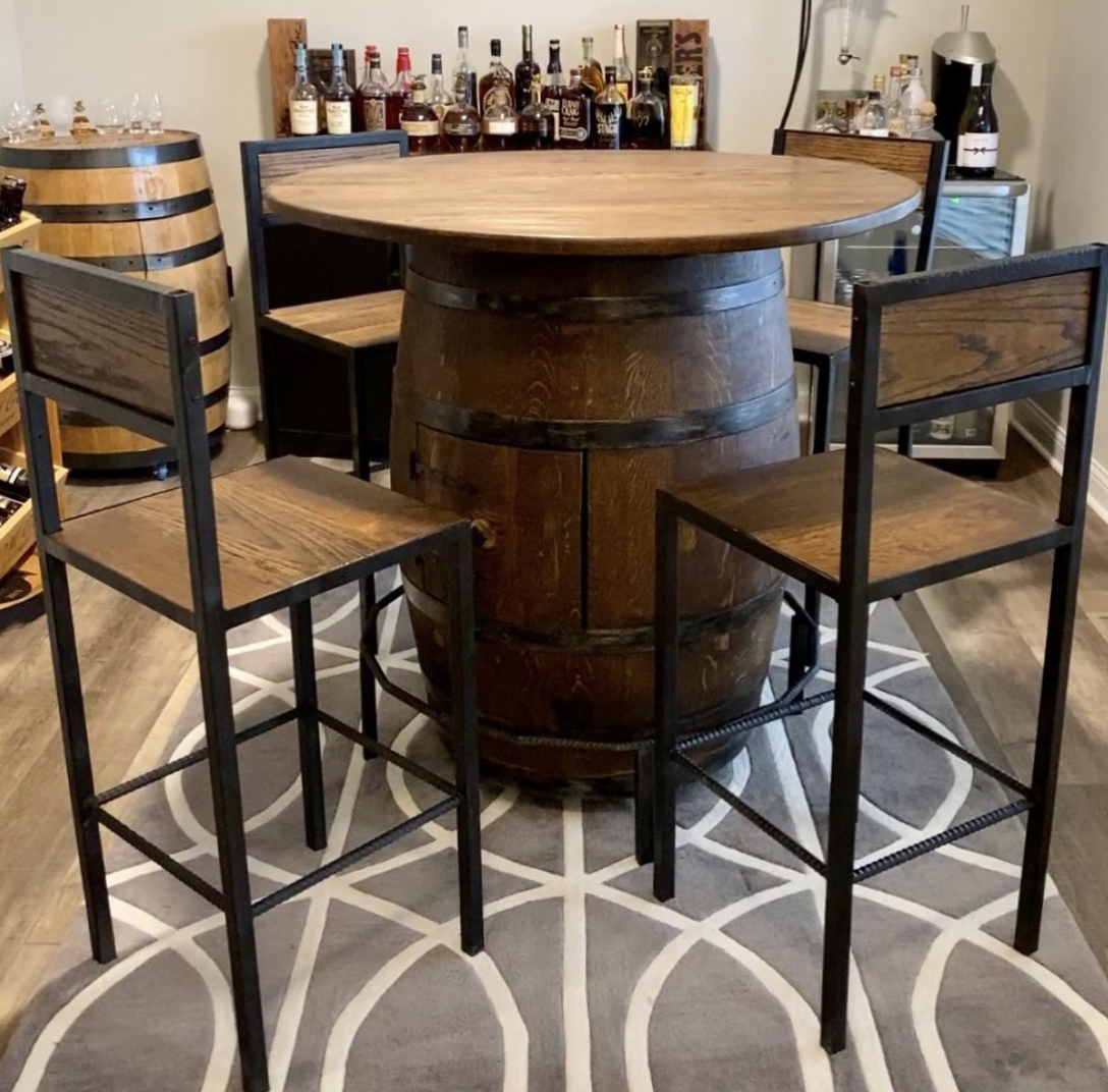 A round wooden table made from a wine barrel with four matching wooden chairs with metal frames around it. In the background, there is a shelf with various bottles of alcohol, a wine barrel, and a small mini fridge.