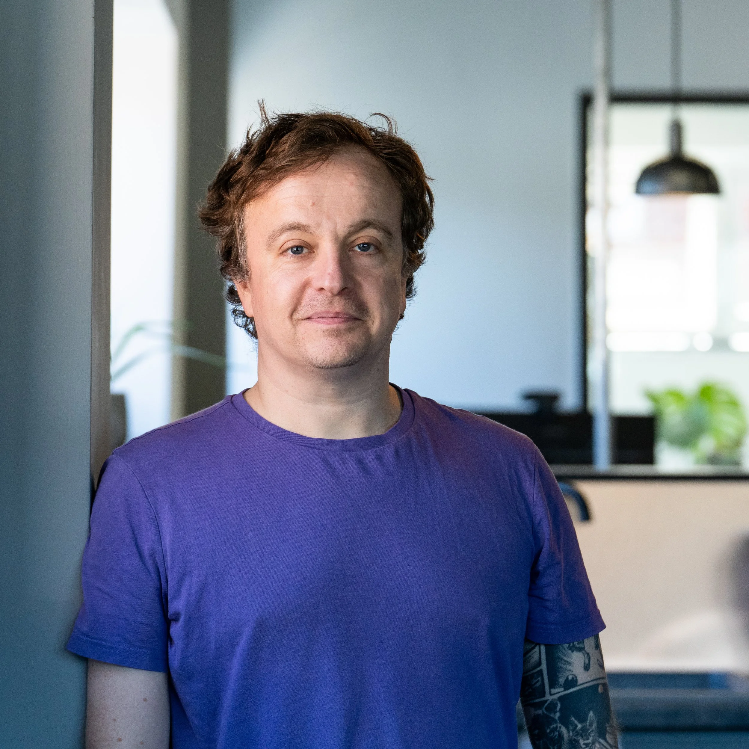A man with light skin, curly brown hair, wearing a purple T-shirt, standing indoors near a gray wall, with a modern office background.