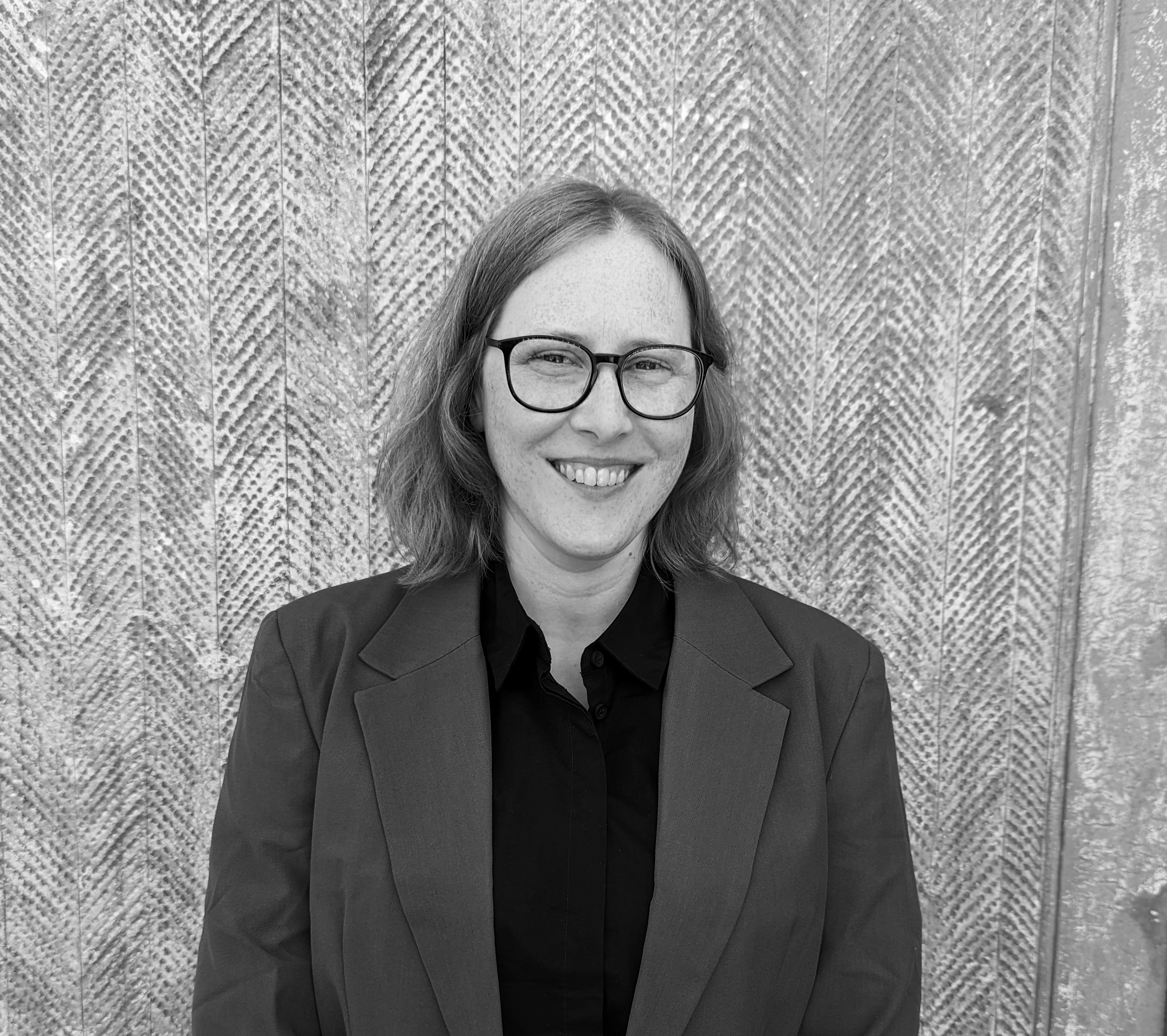 A woman with shoulder-length hair, glasses, and a suit, smiling in front of a textured wall.