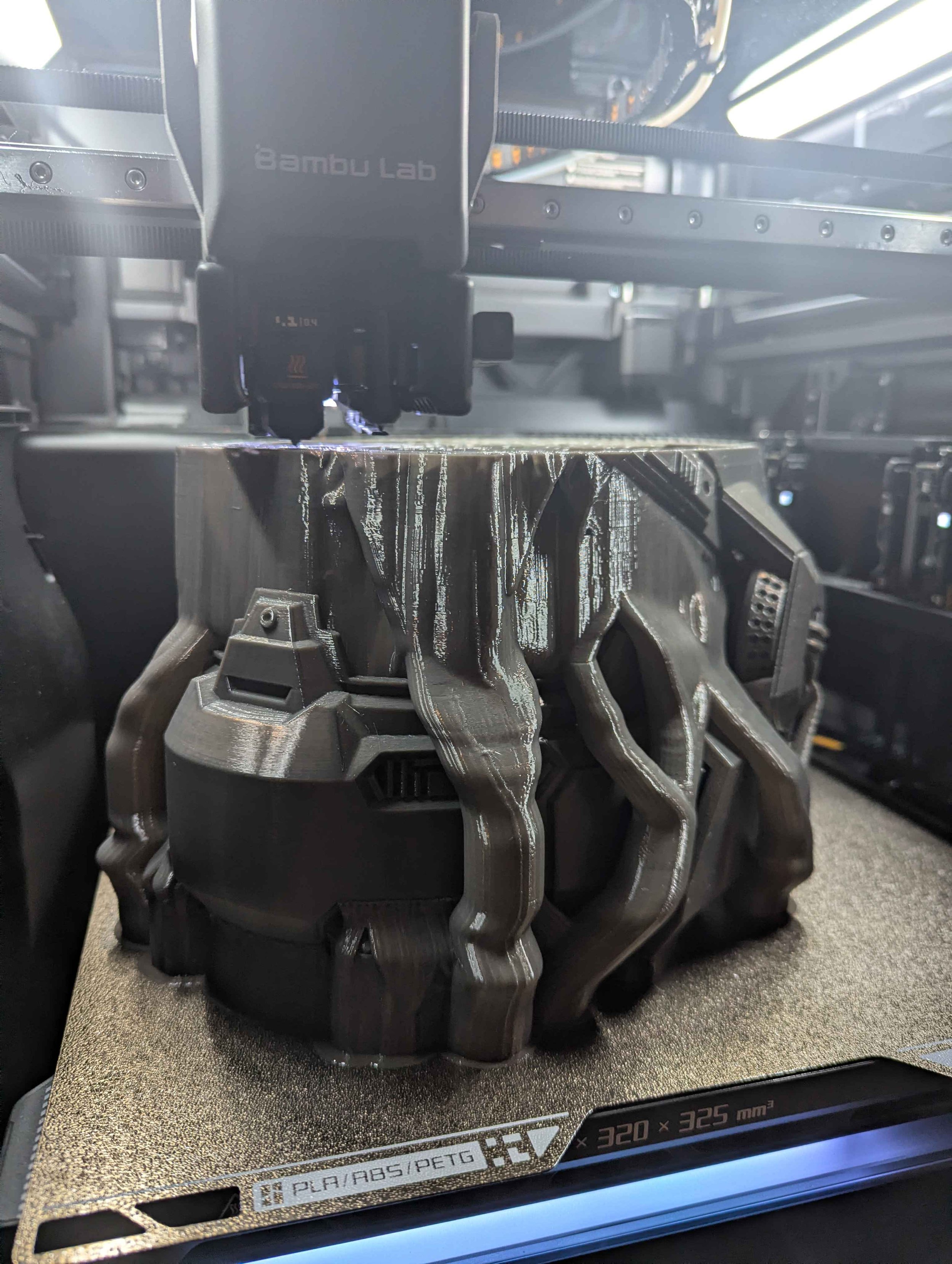 3D-Drucker im Einsatz, der eine schwarze, komplexe, strukturierte Kunststofffigur druckt, mit sichtbarem Druckkopf und Druckbett.