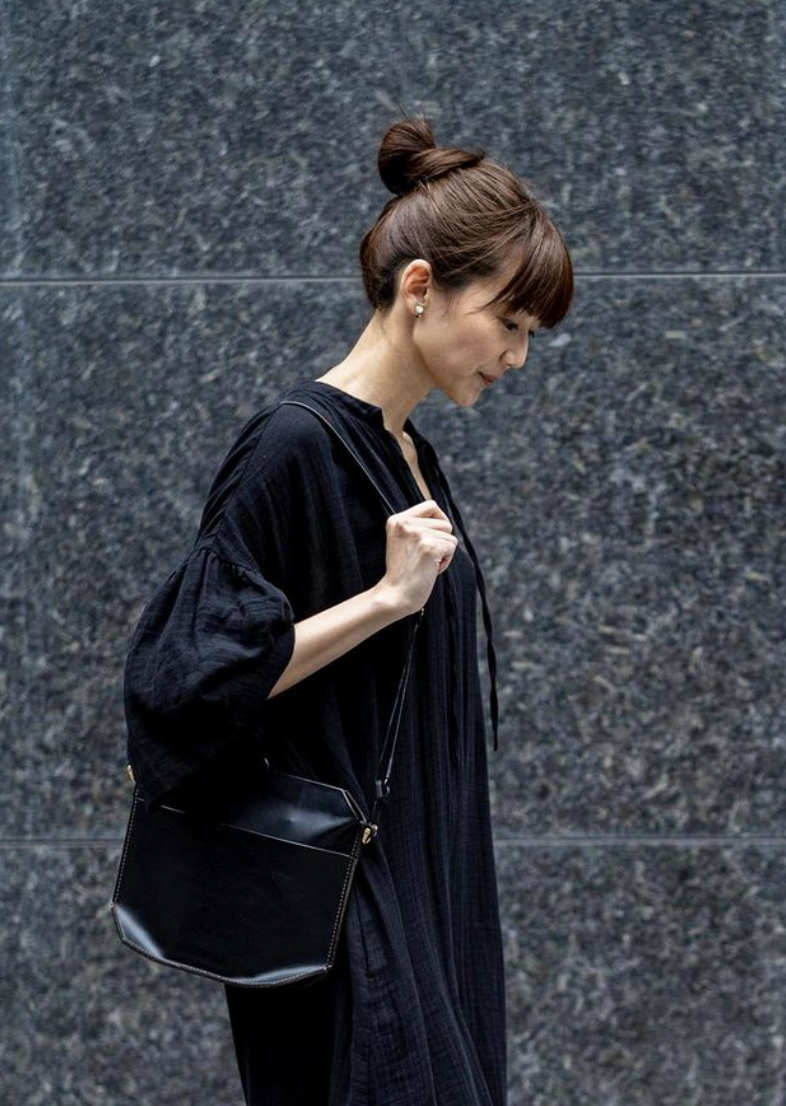 Side profile of a woman with brown hair in a bun, wearing a black dress and earrings, carrying a black purse, standing against a dark, textured wall.