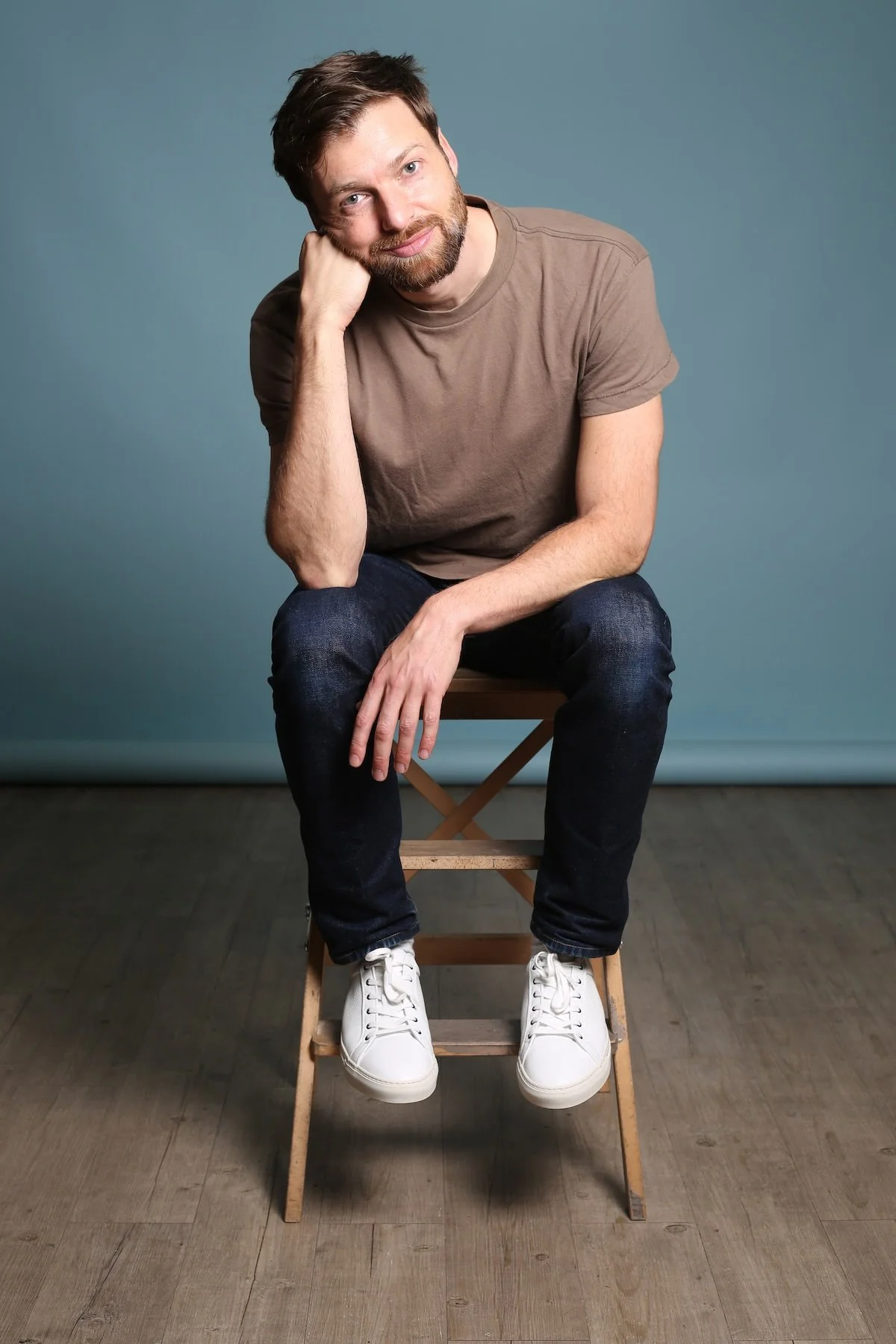 Stéphane Vernier assis sur une petite échelle en bois, portant un t-shirt beige, un jean foncé et des baskets blanches, posé contre un fond bleu