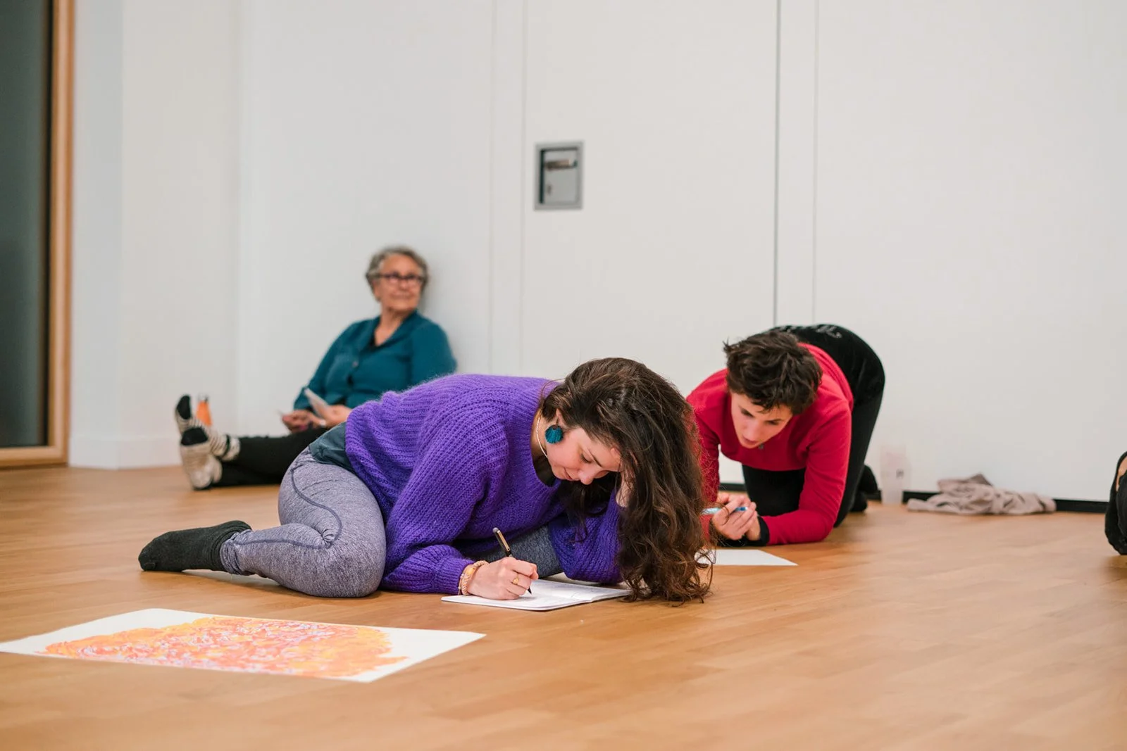 Deux femmes à genoux sur un sol en bois, en train d'écrire, dans une pièce à murs blancs. Une femme plus âgée est assise contre le mur en arrière-plan, regardant vers eux, lors des dimanches Life Art Process® de Stéphane Vernier.
