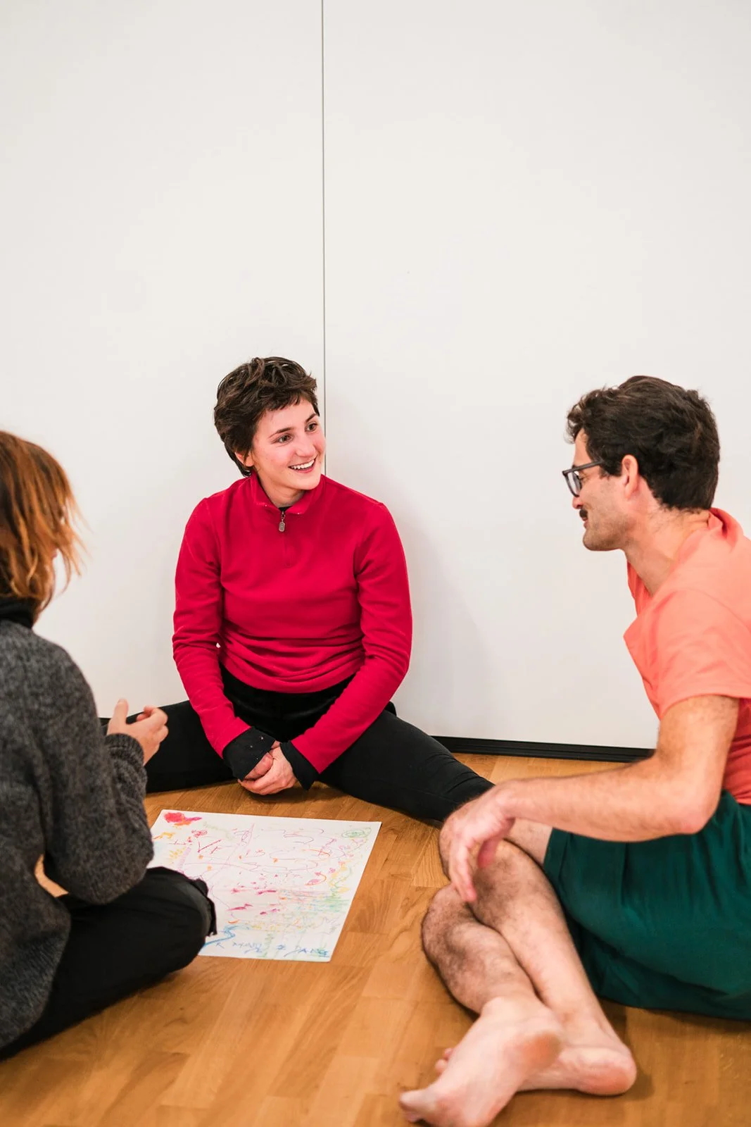 Trois personnes assises par terre dans une pièce claire, discutant et souriant, avec un dessin coloré sur la feuille devant elles, lors des dimanches Life Art Process® de Stéphane Vernier.