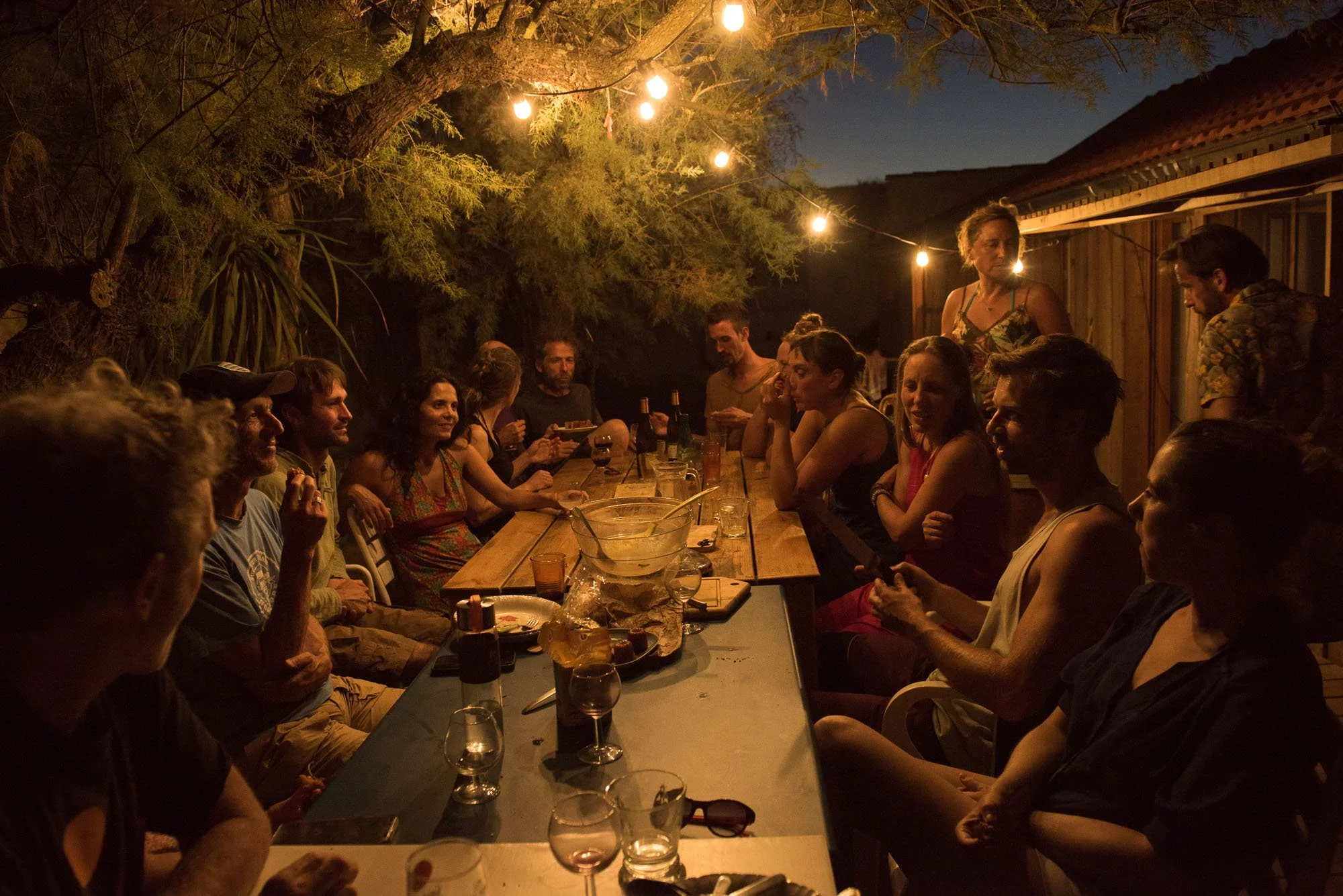 Groupe de personnes réunies autour d'une table lors d'une fête en soirée, éclairée par des guirlandes lumineuses, en plein air avec un ciel nocturne, lors du stage La Sagesse des Vagues.