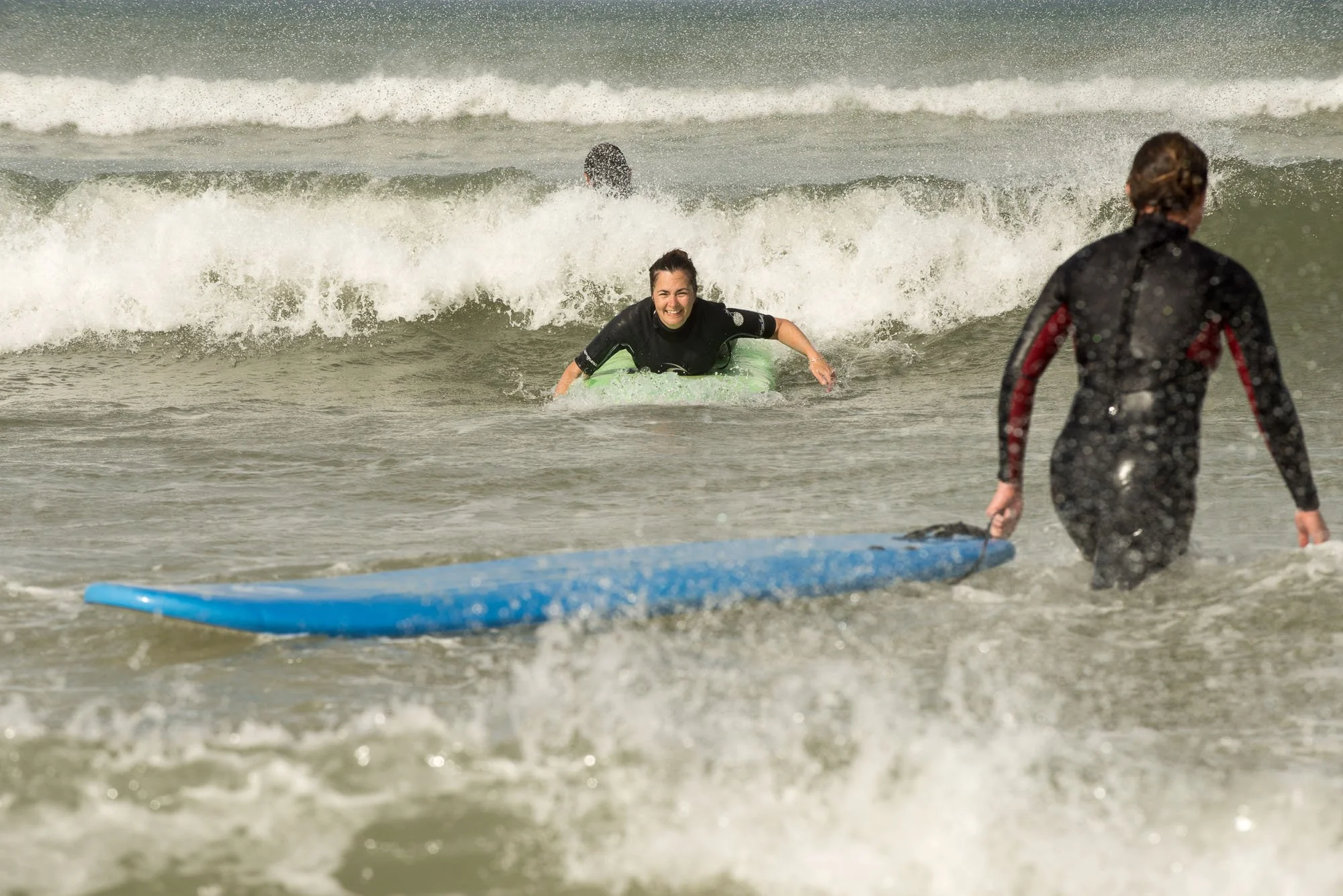 Trois personnes en train de faire du surf dans l'océan, deux en position sur leur planche et la troisième marchant dans l'eau avec une planche bleue, lors du stage La Sagesse des Vagues.