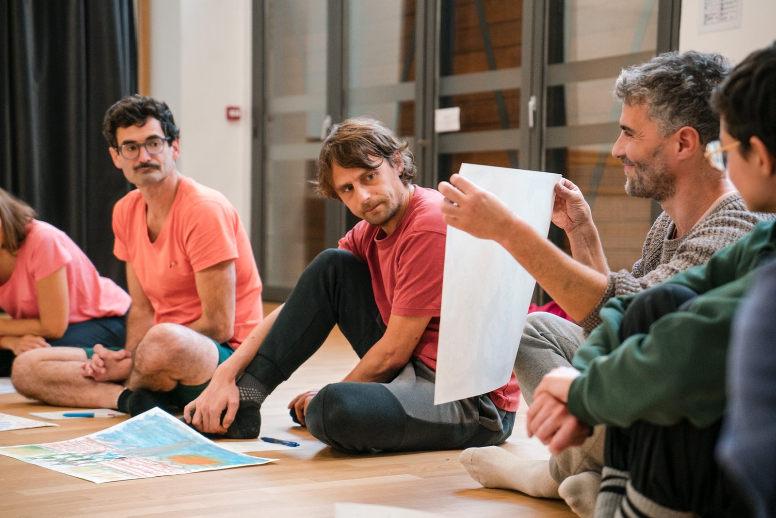 Groupe de personnes assises en cercle sur un sol en bois, discutant lors d'un atelier Life Art Process®, avec des dessins devant eux.