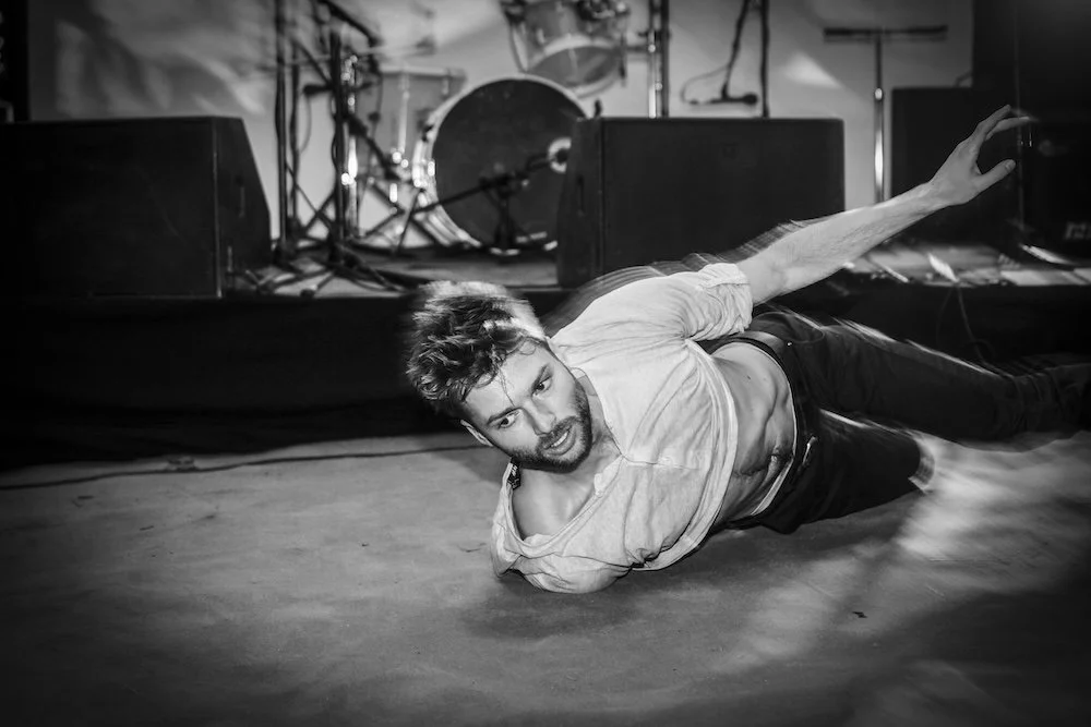 Stéphane à la chevelure en désordre, allongé sur le sol d'une scène de concert, en pleine performance, avec des instruments de musique en arrière-plan.