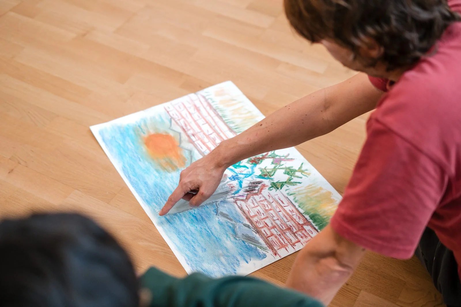 Une personne pointe un dessin ou une peinture à plat sur le sol en bois, représentant une scène avec un soleil, de l'eau, des montagnes, et une clôture en brique, lors des dimanches Life Art Process® de Stéphane Vernier.