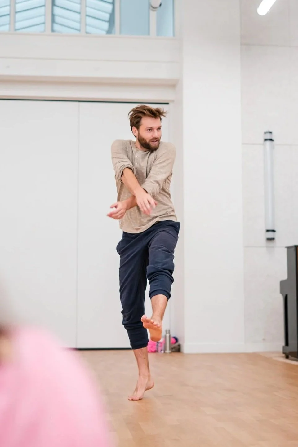 Stéphane Vernier danse dans une salle de danse avec un parquet en bois, en levant une jambe et en croisant les bras, dans un espace lumineux et moderne.