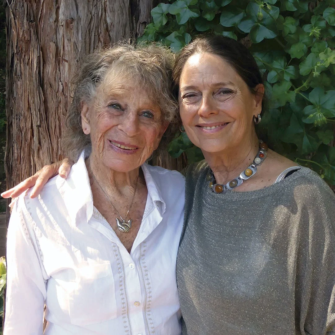 Anna Halprin et Daria Halprin, souriantes posent ensemble près d'un arbre et de feuillage vert.
