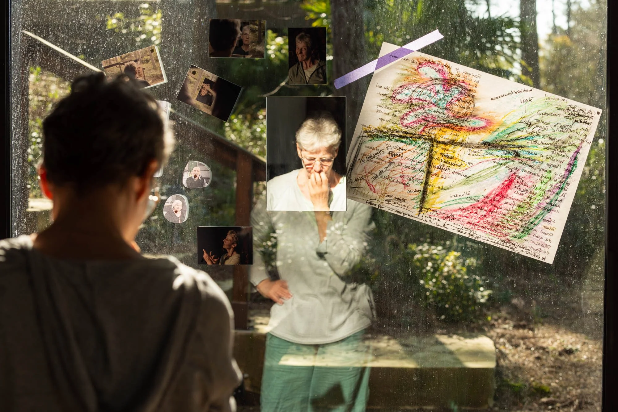 Une femme âgée regarde un tableau d'affichage avec des photos et des dessins, vus à travers une vitre, avec une arrière-plan de jardin ensoleillé, lors du stage Regards animé par Isabelle Chapuis et Stéphane Vernier.