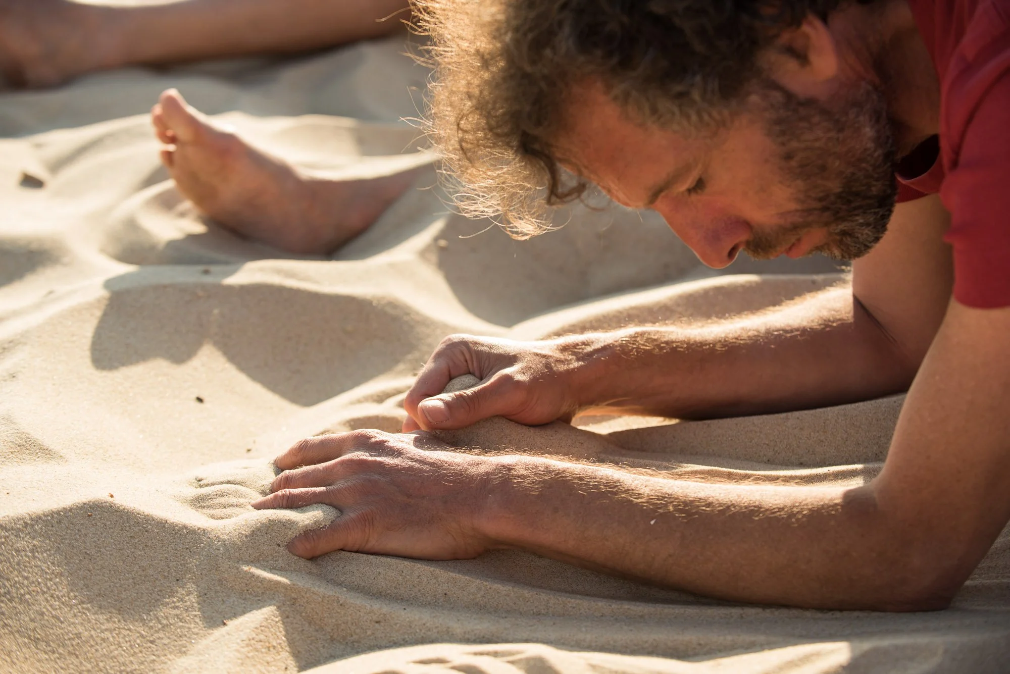 Un homme à la barbe fait de la relaxation ou de la méditation en position couchée sur le sable lors d'une journée ensoleillée, lors du stage La Sagesse des Vagues.