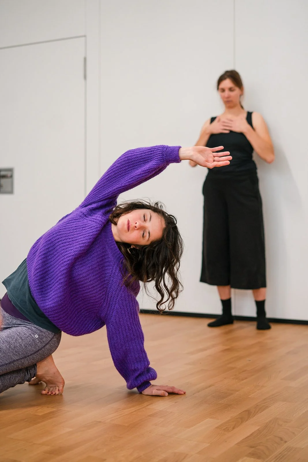 Deux femmes faisant de la danse contemporaine dans une pièce avec sol en bois. La femme au premier plan porte un pull violet et se penche, les yeux fermés, avec une main au sol et l'autre levée. La femme au fond, en robe noire, se tient contre un mur, l'air concentrée.