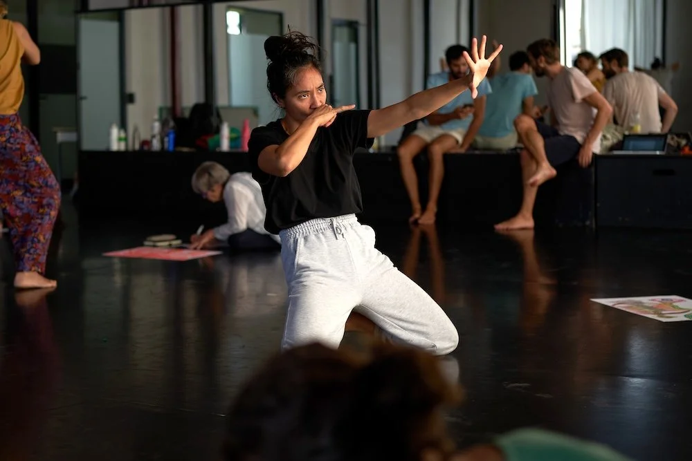 Une jeune femme en tenue décontractée danse dans une salle de danse, entourée de plusieurs autres personnes assises ou debout, en train de discuter ou d'observer, lors d'un atelier Life Art Process®.